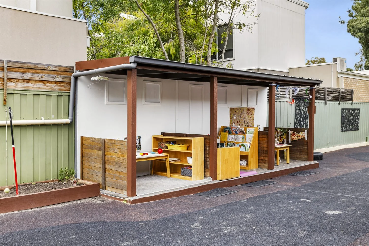 Outdoor classroom with wooden furniture and educational materials, surrounded by a fence, with a small garden bed nearby.