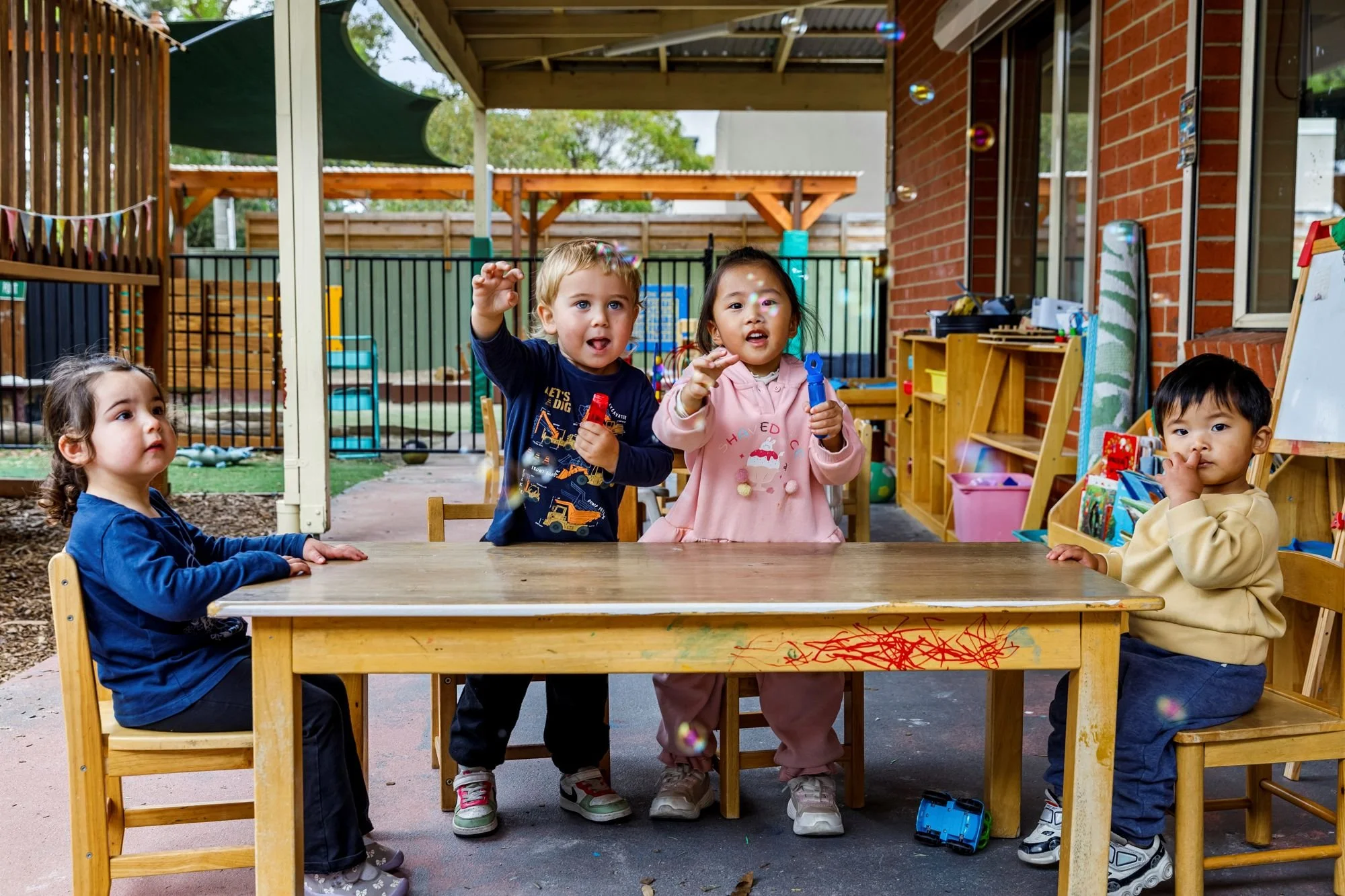 Children sitting at a wooden table in an outdoor play area, with some children reaching for bubbles.