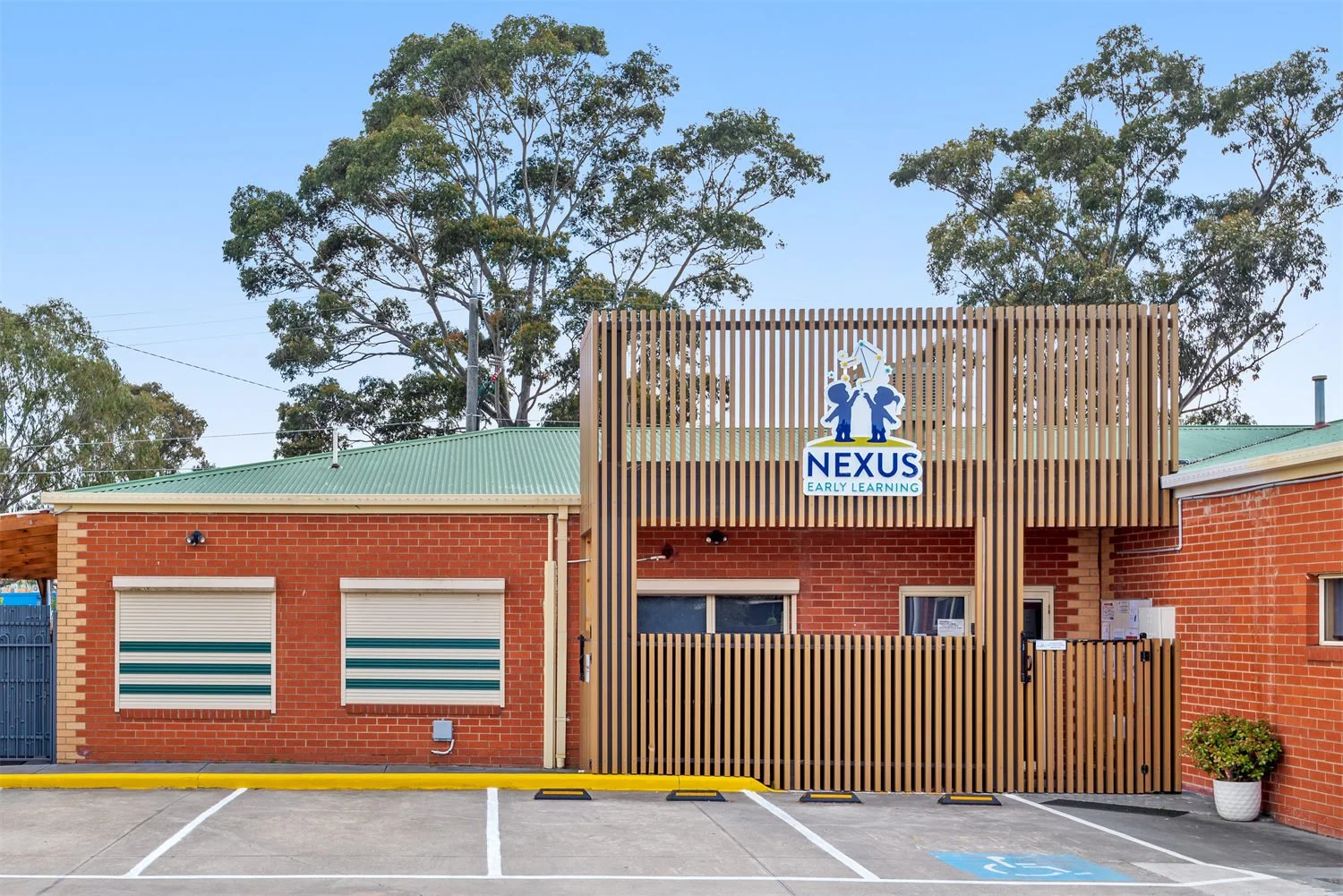 Red brick building with a green metal roof, wooden slat fencing, and a sign reading "NEXUS Early Learning" above the entrance. Parking lot in front with marked spaces, including one for handicapped parking, and trees in the background.