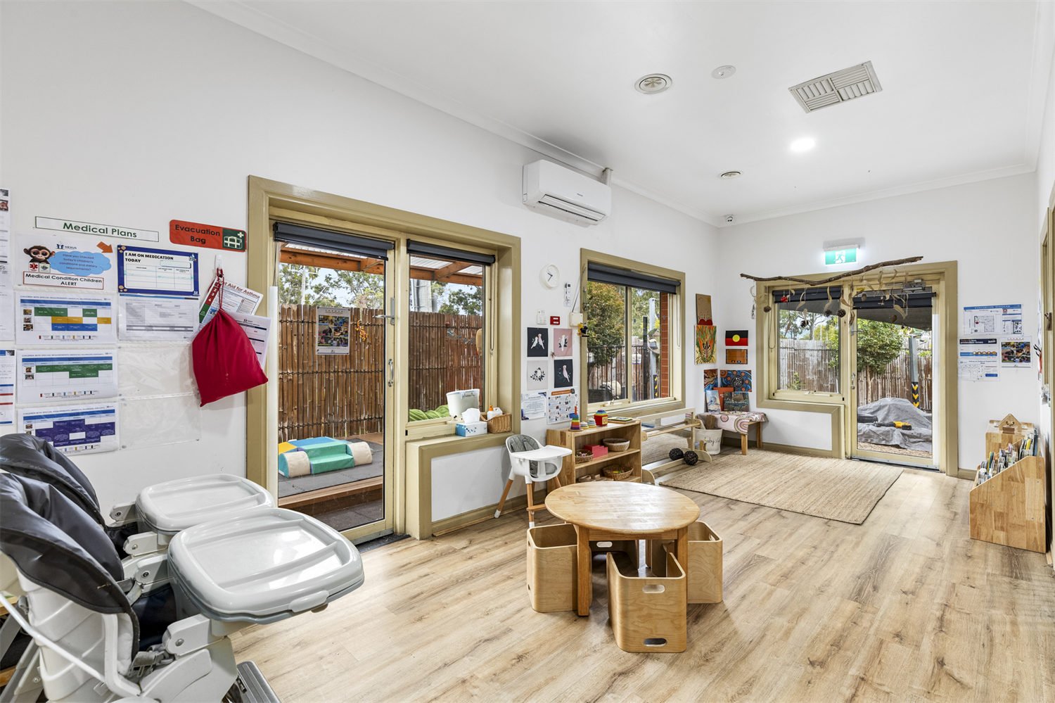 Bright classroom with windows, children's toys, art, and seating, featuring a white wall with posters, a wooden table with chairs, and an outdoor area visible through sliding doors.