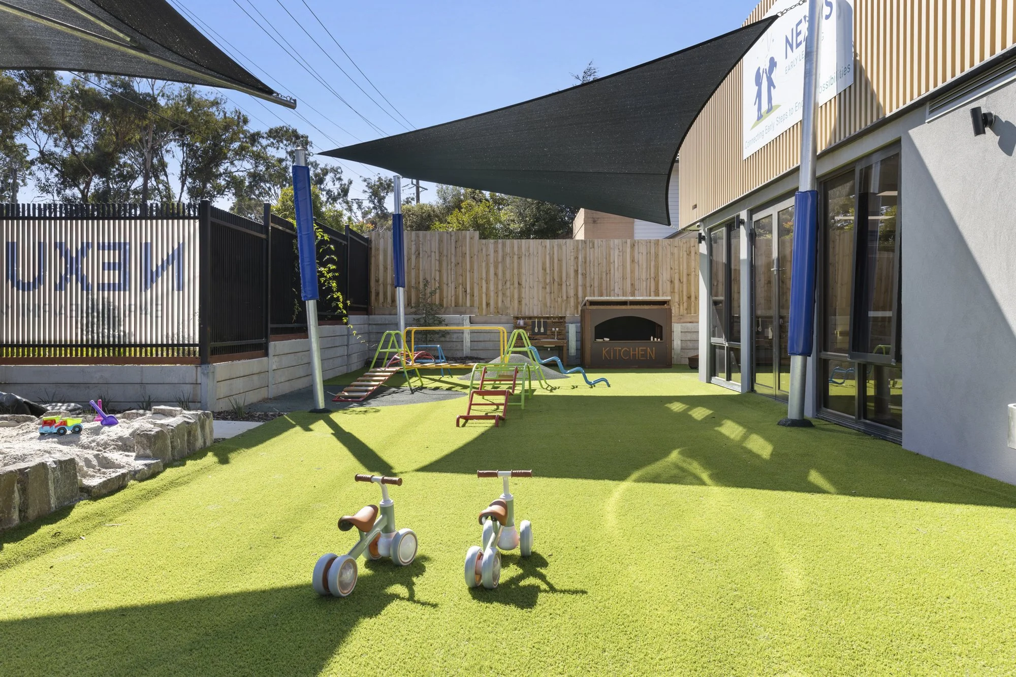 A colorful playground with a grassy area, small balance bikes, a sandpit with toys, a picnic table, shade sails, and a wooden fence, adjacent to a building with large sliding doors and a sign about early learning.