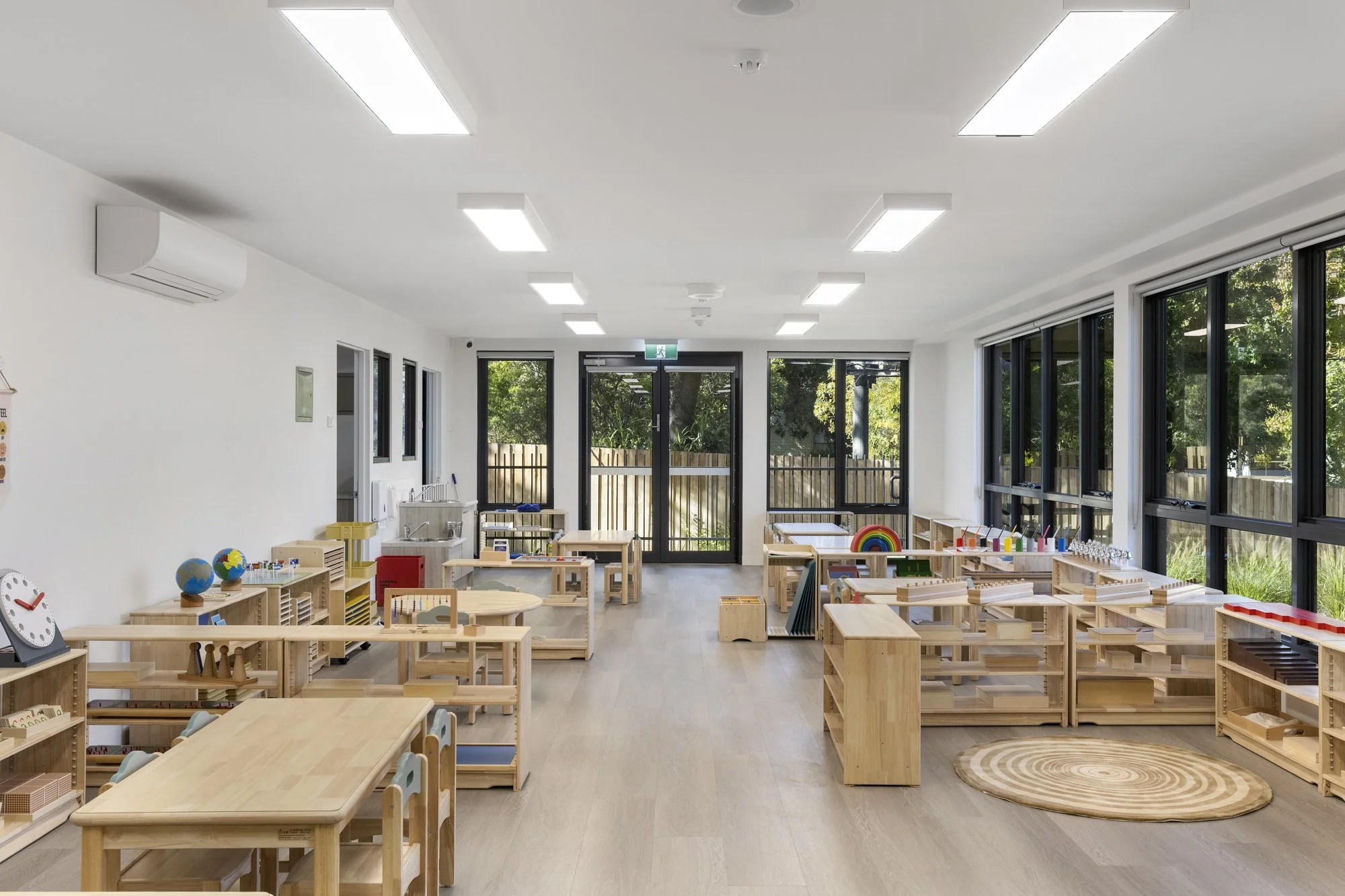 Empty classroom with wooden furniture, educational toys, and large windows showing outside greenery.