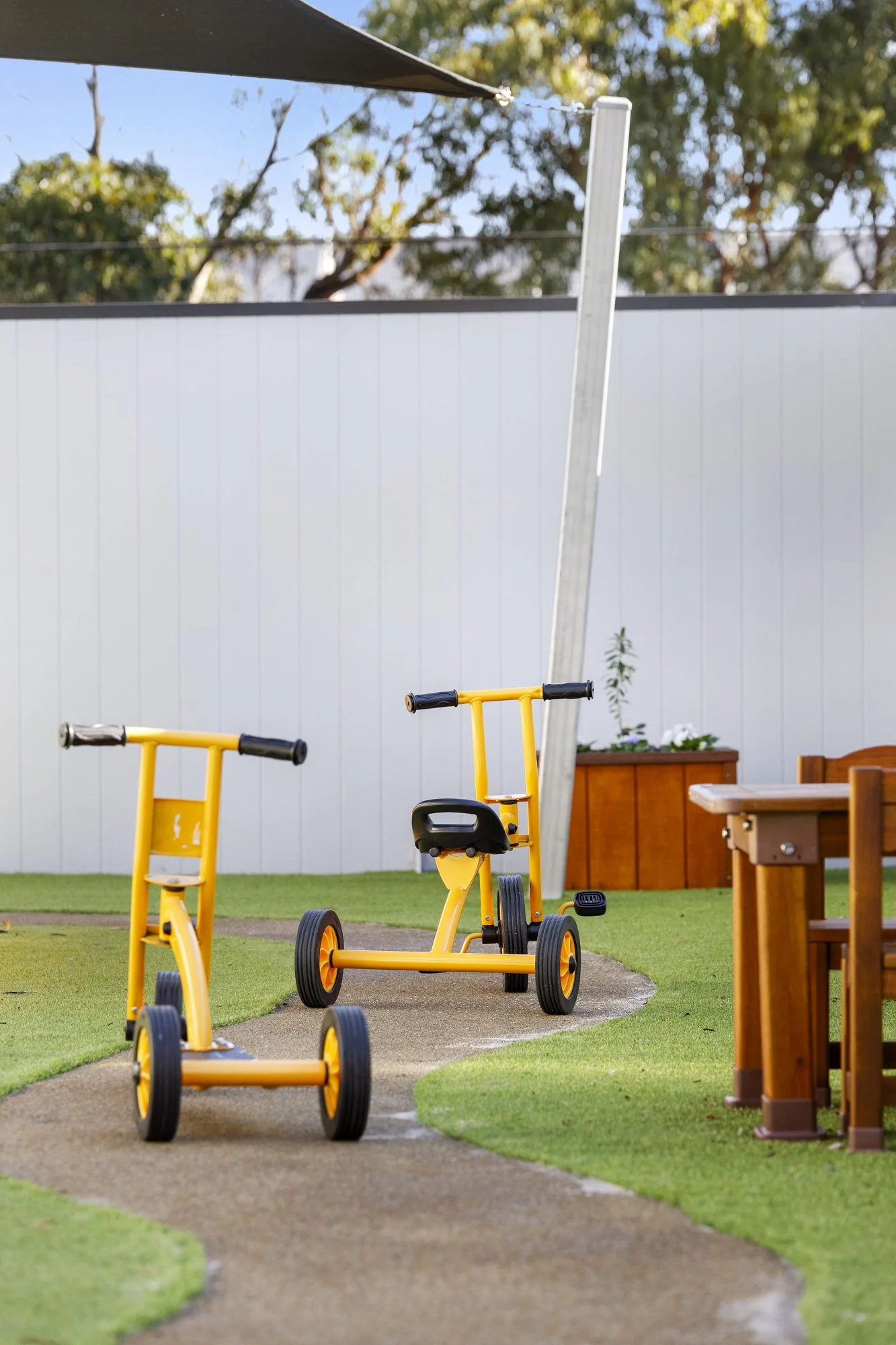 Children's outdoor playground with yellow tricycles on a winding path, wooden benches, planters, and a white fence under a sunny sky.
