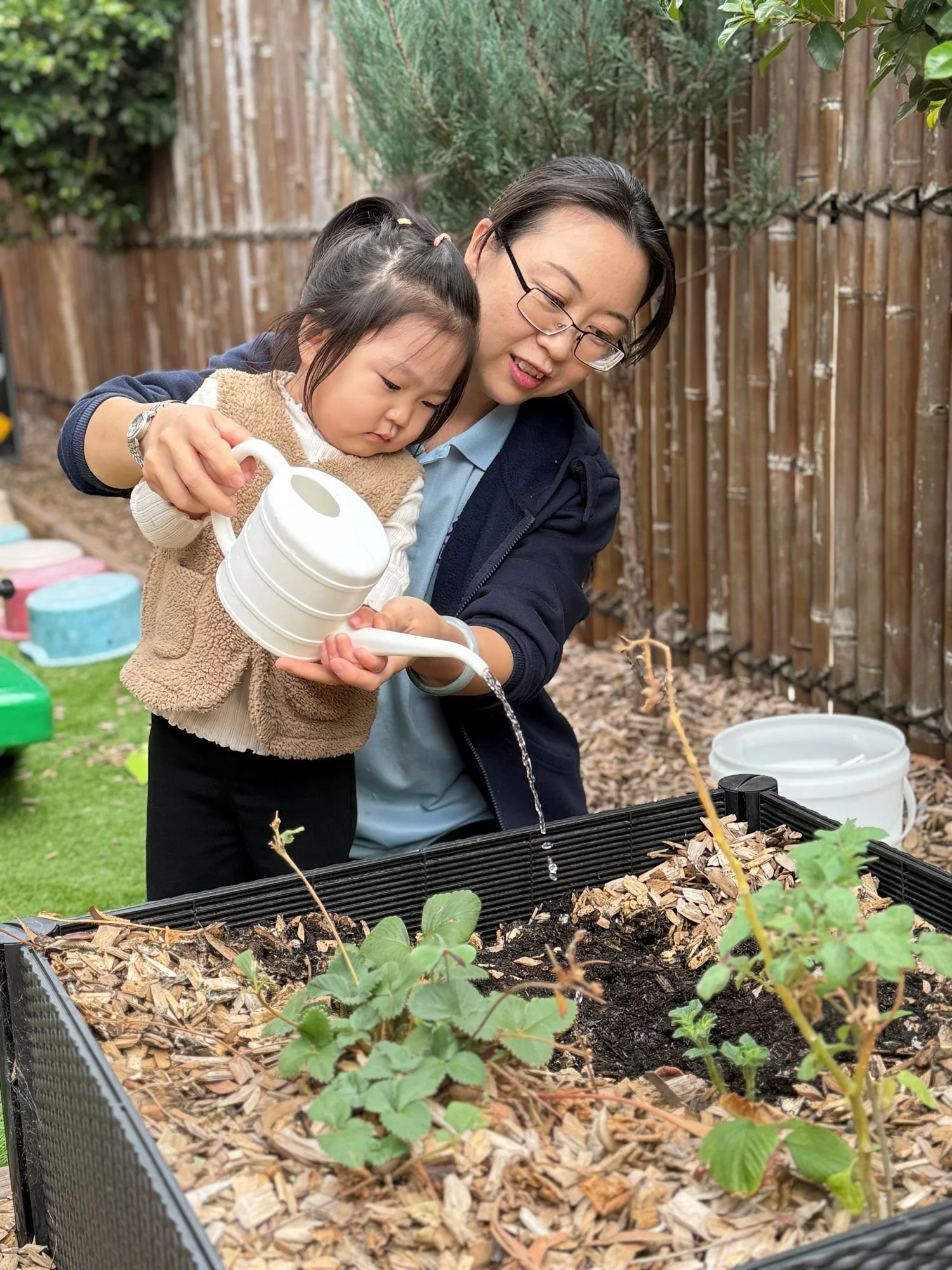 A woman and a young girl planting seedlings in a garden bed outdoors, with tools and buckets nearby, a wooden fence in the background.