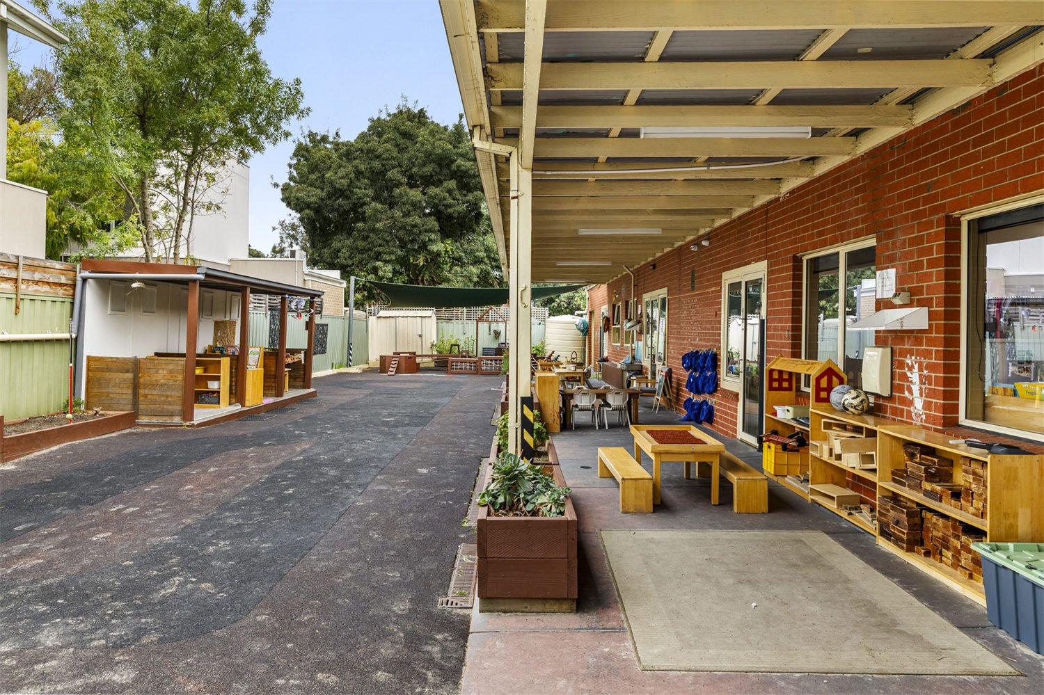 Outdoor area of a school or daycare with a covered porch, playground equipment, and outdoor furniture, surrounded by a fence and trees.