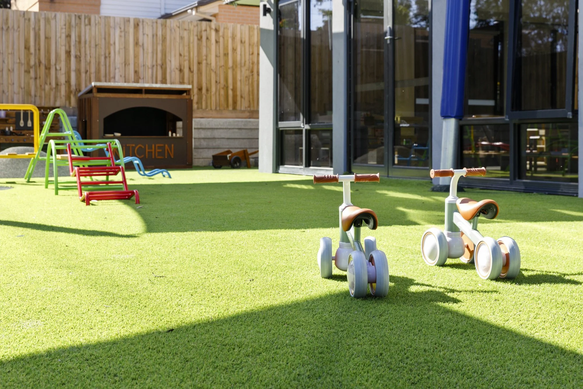 A backyard play area with a green artificial turf surface, colorful plastic swings, a tricycle, a wooden cart, and a wooden playhouse. There is a modern glass house with sliding doors and a fence in the background.