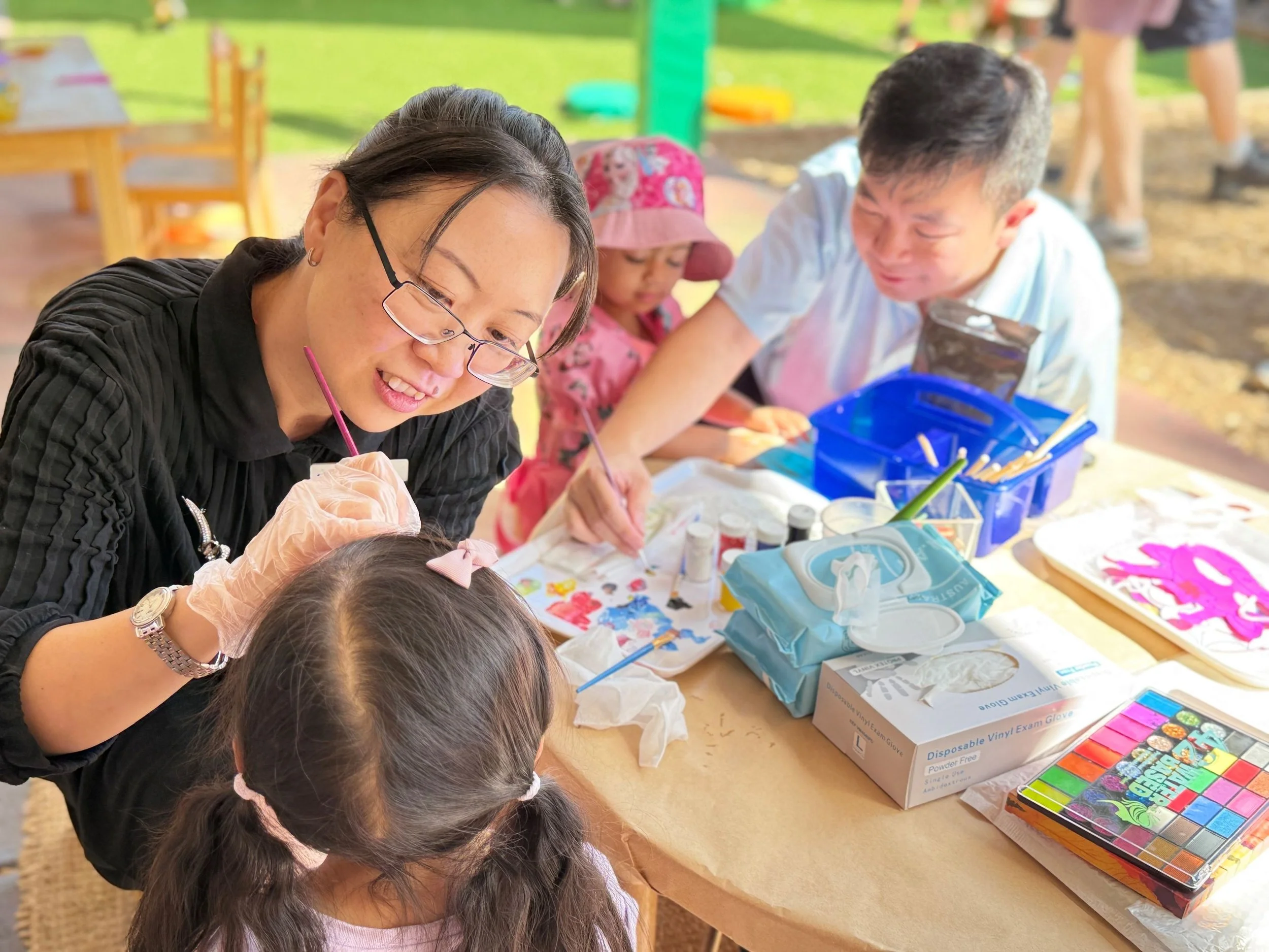 People participating in a crafts activity outdoors, with art supplies like paints, brushes, and paper on a table.