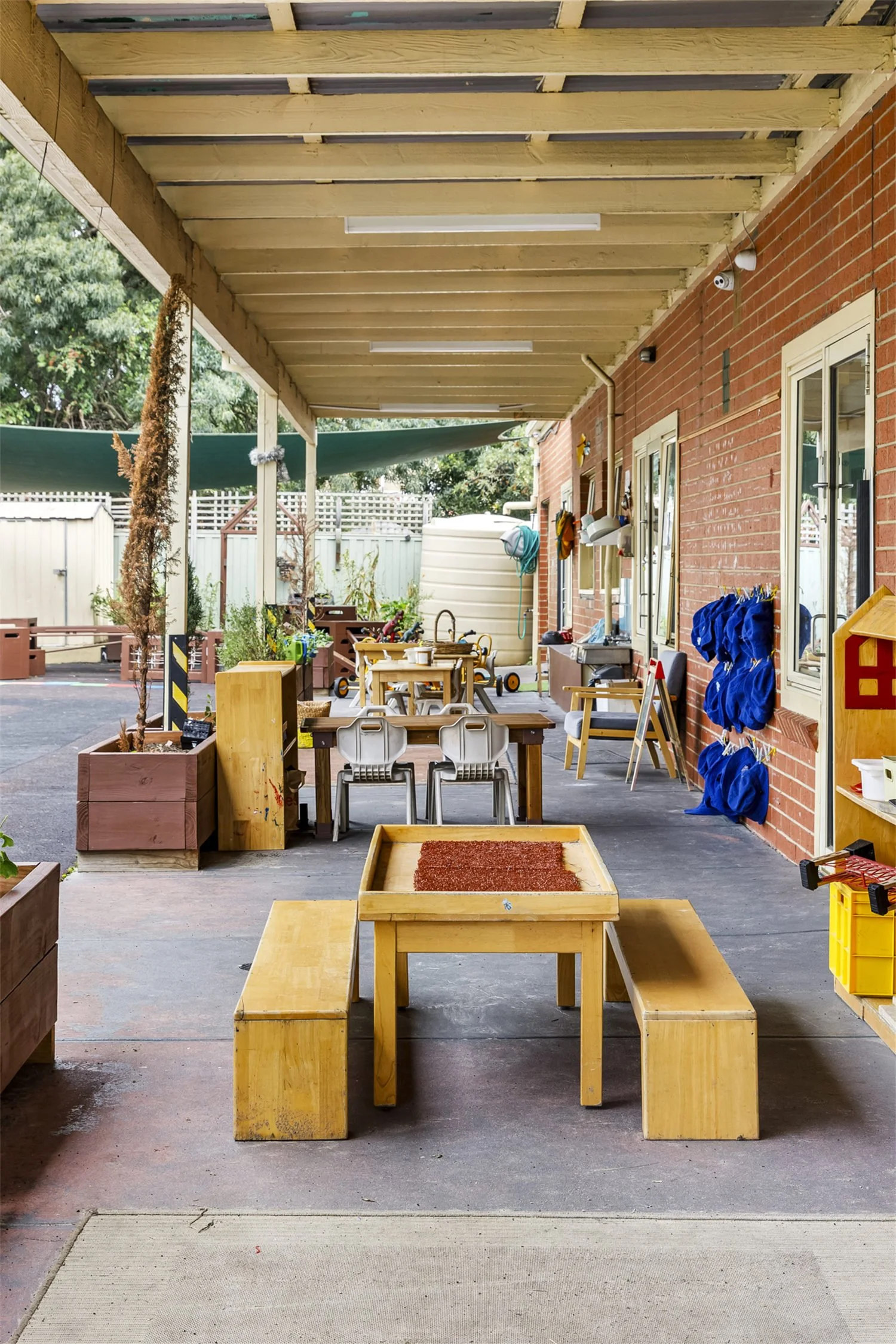 View of an outdoor covered area attached to a brick building, with children's tables and chairs, toys, plants, and various items hanging on the wall, and a shed and trees visible in the background.