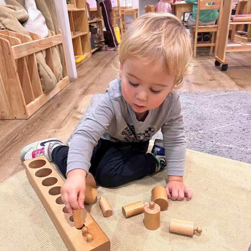 Young boy playing with wooden peg puzzle pieces on a beige carpet in a classroom.