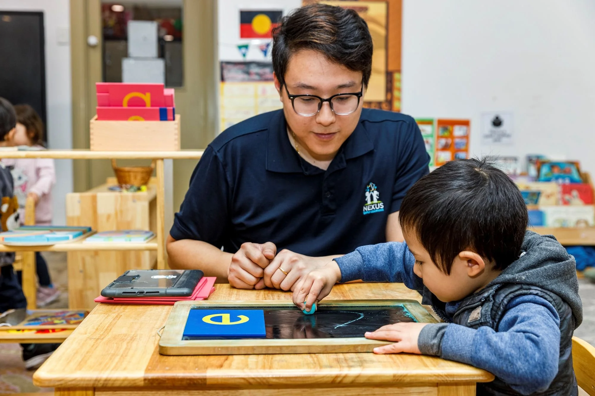 A man wearing glasses and a black shirt with a Nexus logo helps a young boy with a tablet at a wooden table in a classroom. Other children are visible in the background, and classroom decorations are on the walls.