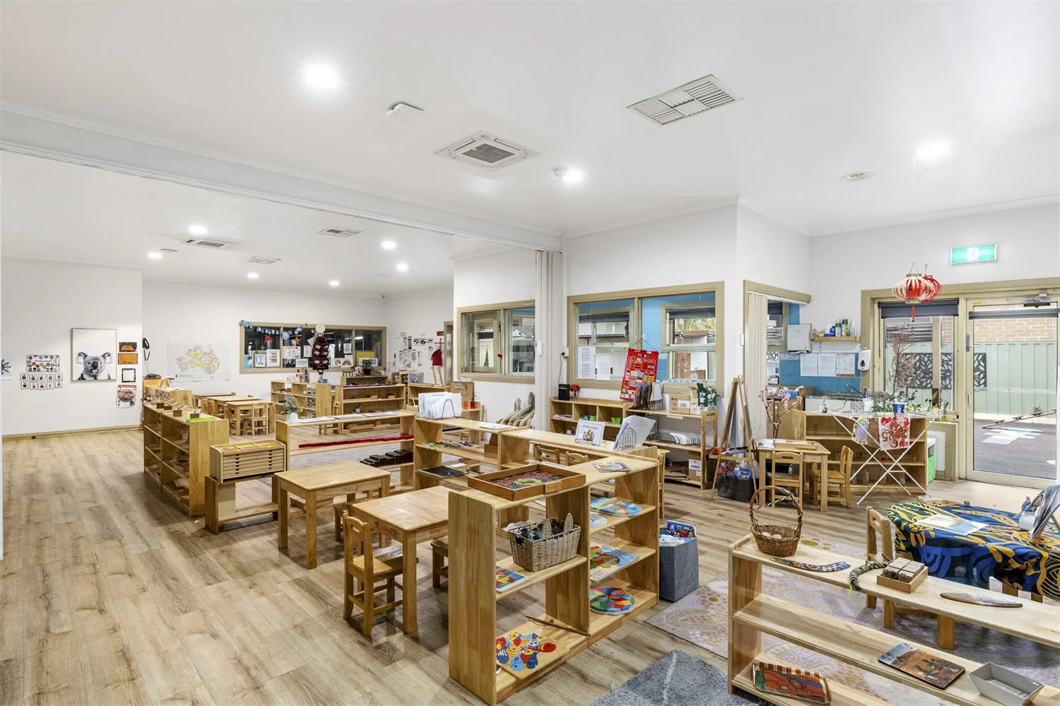 A classroom or daycare room with wooden furniture, shelves with toys and books, and windows letting in natural light.