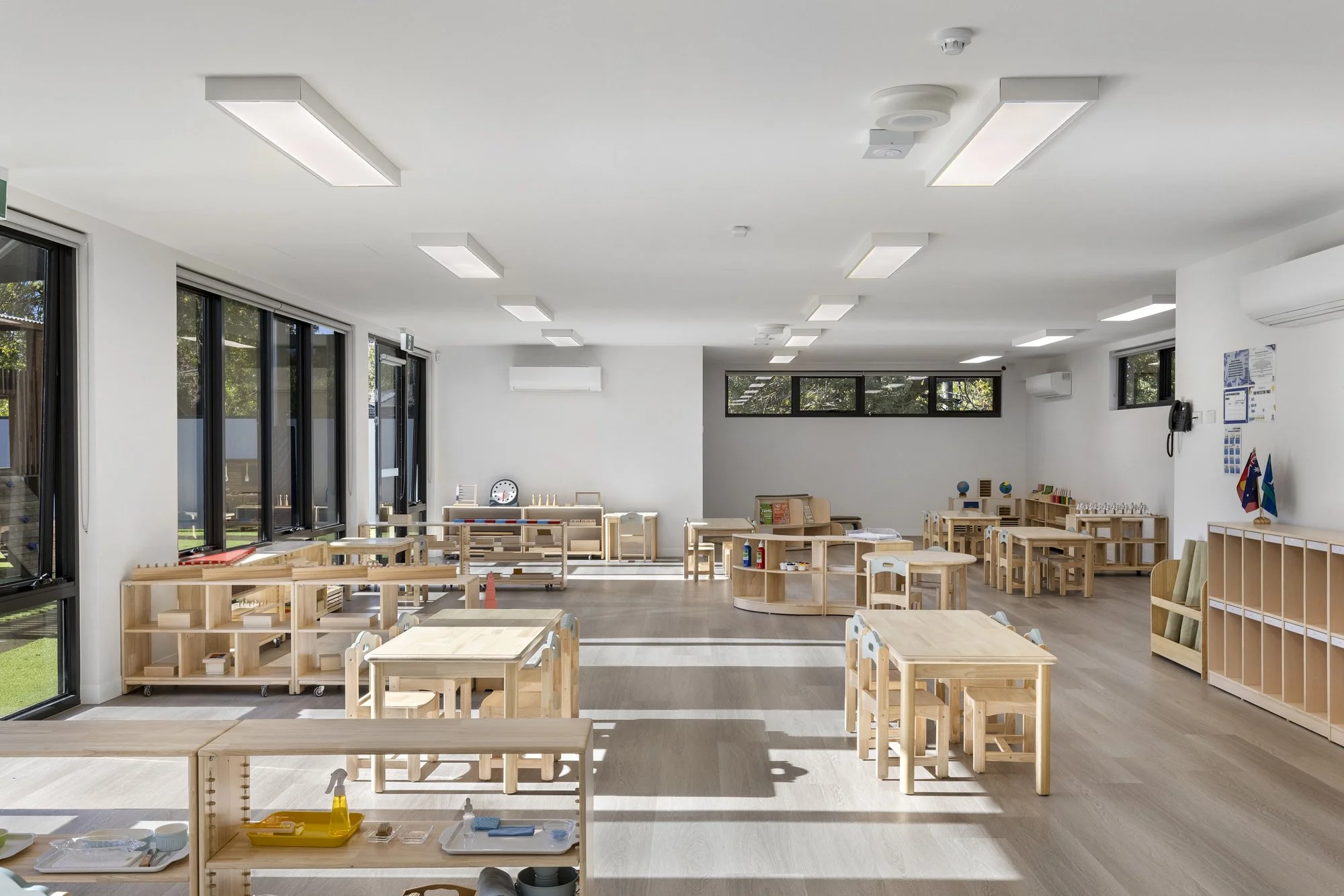An empty modern classroom with large windows, wooden tables, and educational materials for young children.
