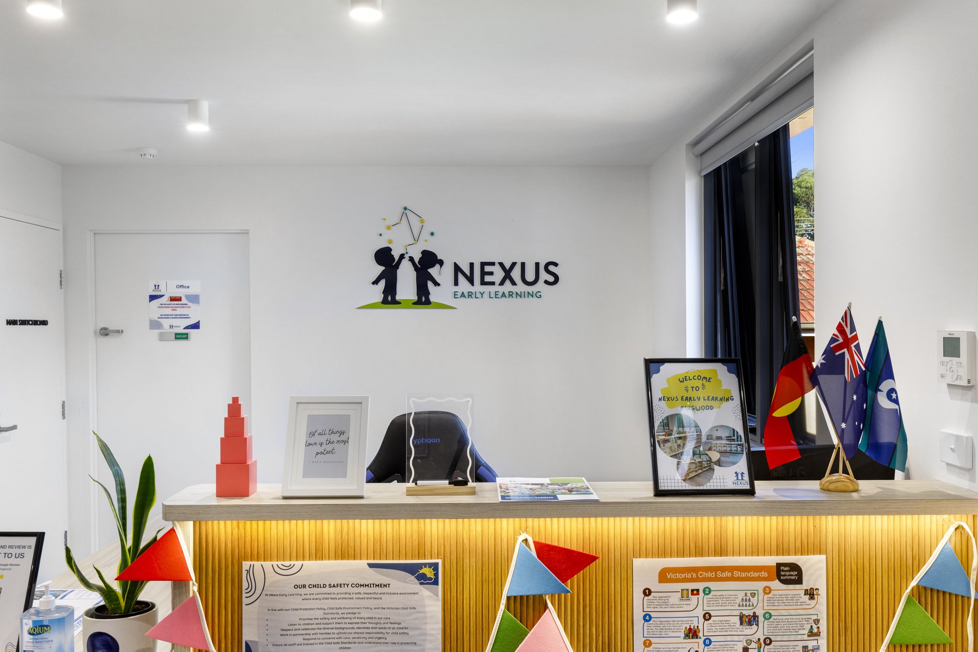 Reception area with a white wall featuring a logo of two children and the words 'NEXUS EARLY LEARNING'. There are flags, framed posters, a desk with a sofa, decorative plants, and classroom supplies. Bright lighting and a window with blue curtains ar
