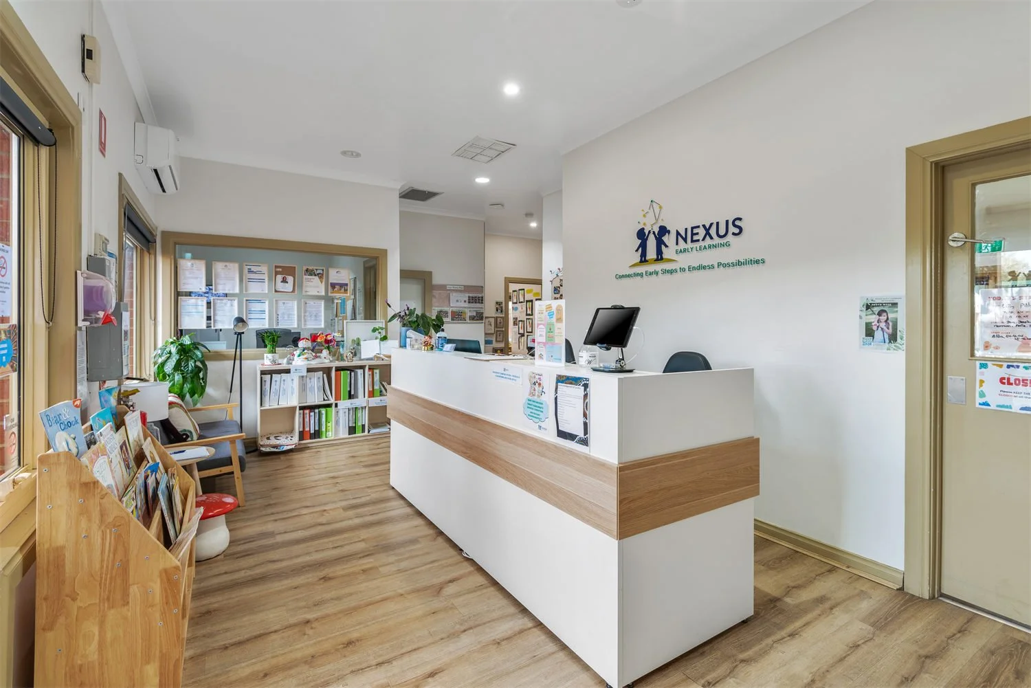 Reception area of a learning center with a white counter, computer, and logo on the wall that reads 'Nexus Early Learning'. There are chairs and bookshelves with children's books, plants, and informational posters.