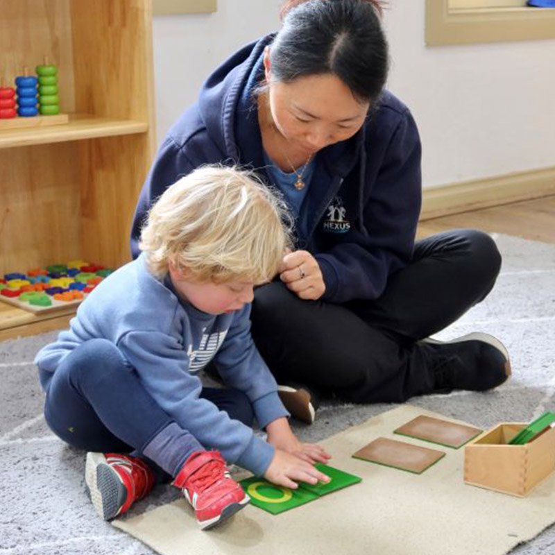 A woman and a young boy sitting on a gray carpet in a room, playing with alphabet tiles. The woman is wearing a navy hoodie and the boy is wearing a gray sweatshirt with red sneakers. There are colorful toys on a wooden shelf behind them.