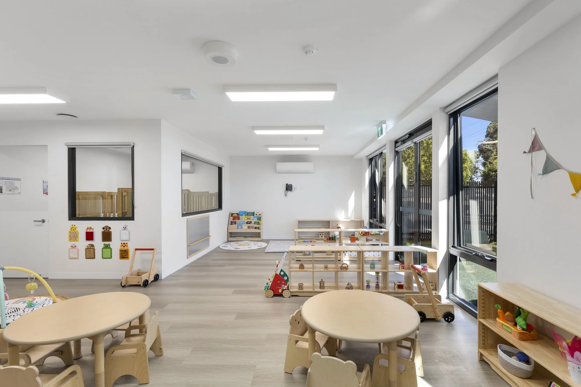 An indoor children's playroom or classroom with natural light coming through large windows. The room includes round tables with chairs, wooden shelves with toys and books, wall decor, and educational materials. There are also educational posters and 