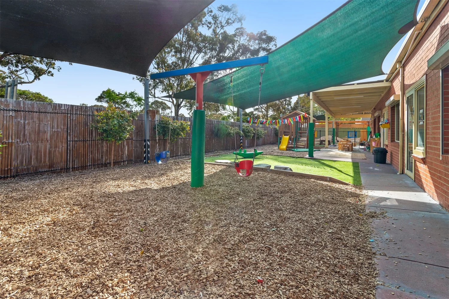 Backyard playground with swings, slide, sandbox, and shaded area, fencing, and concrete walkway along house, sunny day.