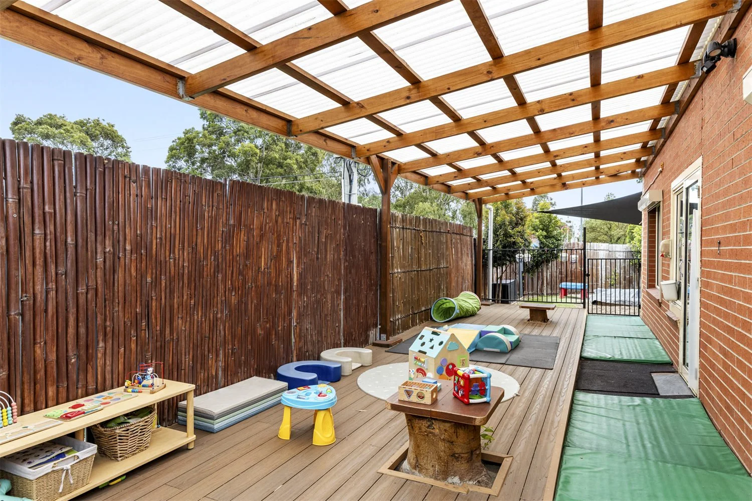 Outdoor backyard with play area for children, including toys, foam mats, tunnel, and low table. Wooden deck and fence, part of a brick house, with green mats along the side and a partially covered roof.