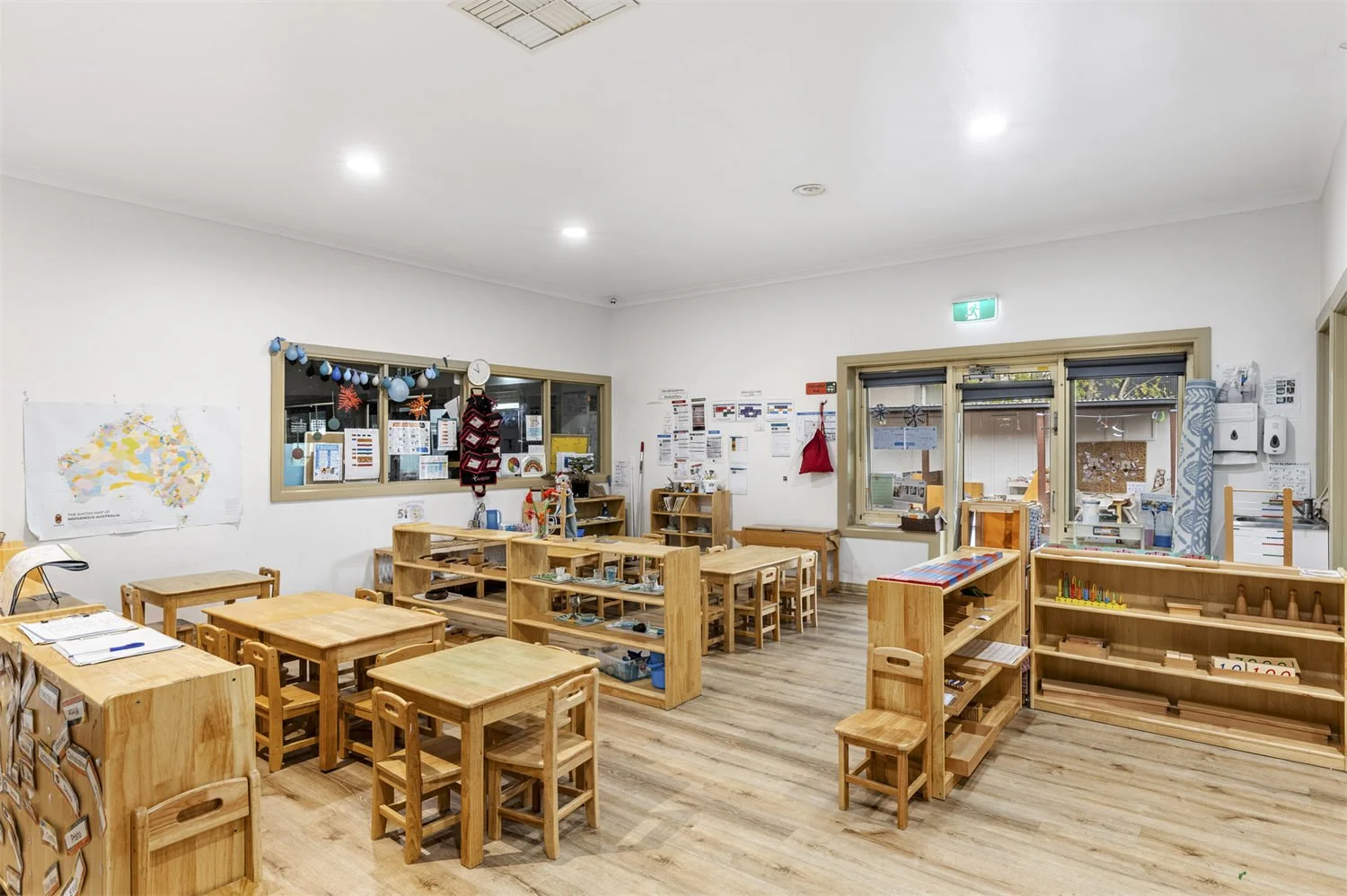 A classroom with wooden tables and chairs, educational materials, and colorful decorations. White walls with windows, posters, and a map. Wooden shelving with supplies and learning tools. Bright lighting and a tidy environment.