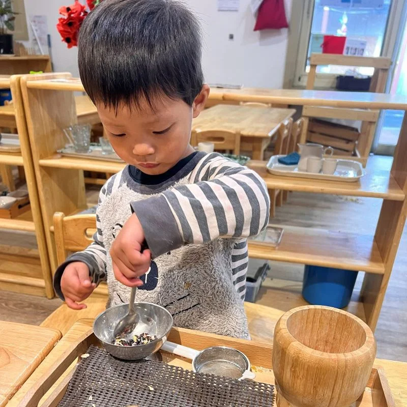A young boy wearing a striped long sleeve shirt scooping colorful dried herbs or tea leaves into a small metal bowl at a wooden table in a classroom or activity room.