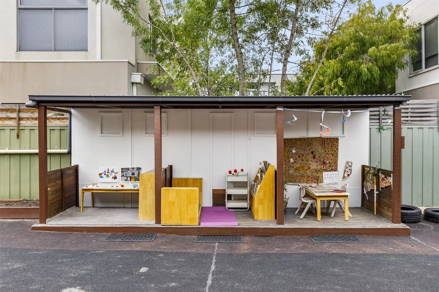 Outdoor classroom with small tables, chairs, and art displays under a covered shelter, surrounded by a green fence and residential buildings, with trees in the background.