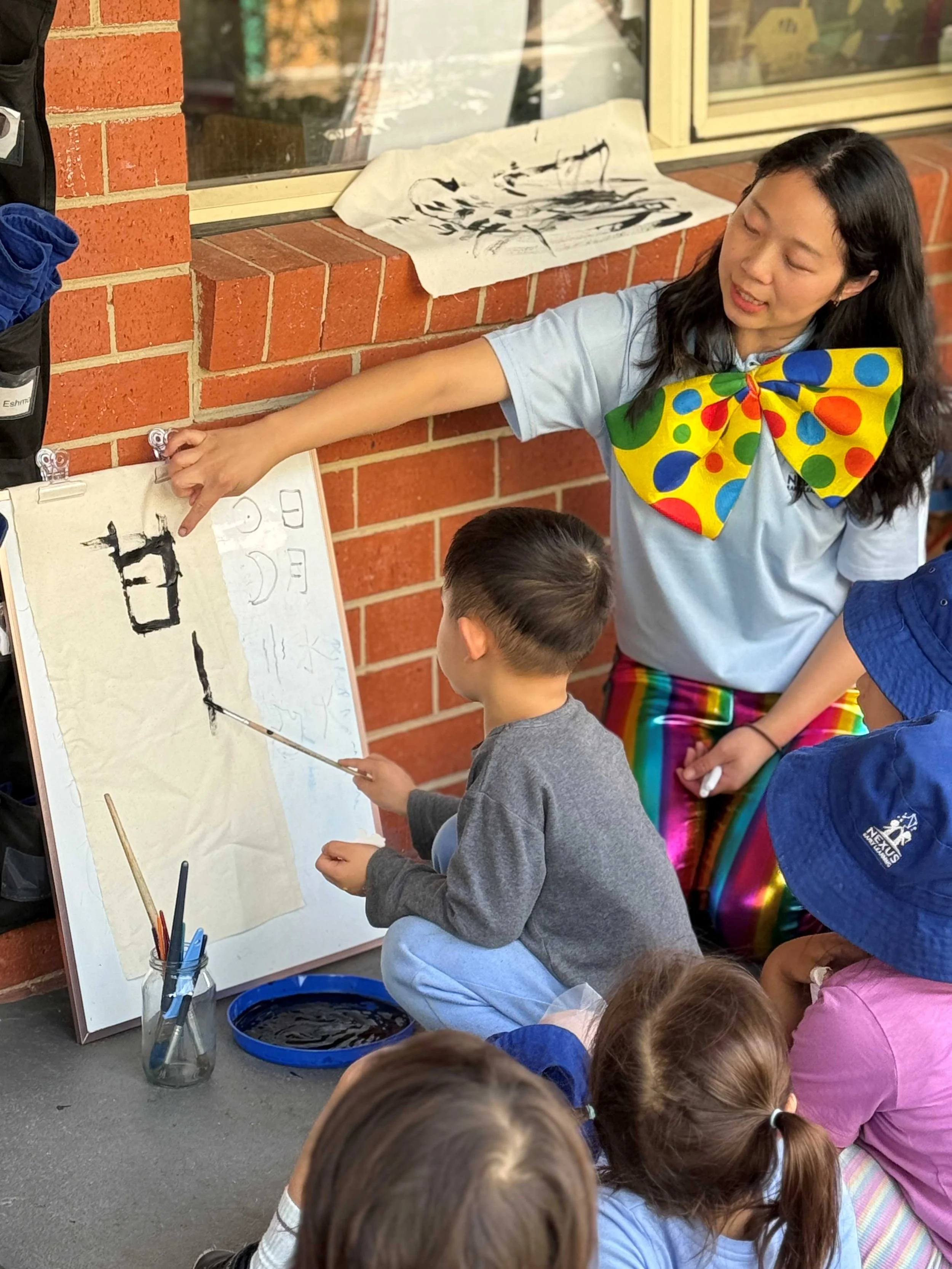 A woman dressed as a clown with colorful polka dot bow ties teaches children watercolor painting outside against a brick wall. She points at a whiteboard with Chinese calligraphy, and children are gathered around watching and holding brushes.