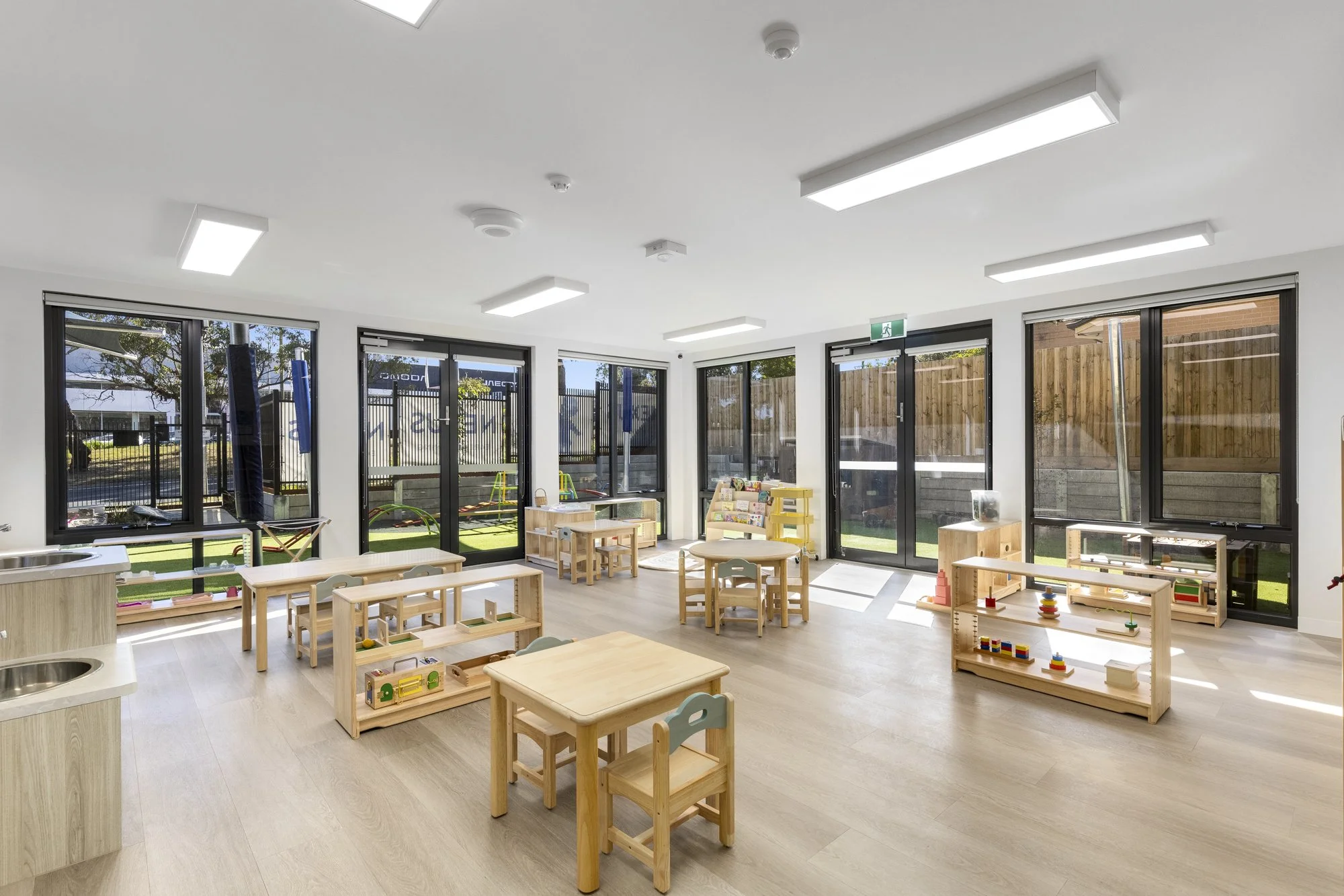 A bright, modern indoor playroom with large windows and sliding glass doors letting in natural light. Small wooden tables and chairs are arranged around the room, which contains various toys and learning materials. Outside, a grassy play area with eq