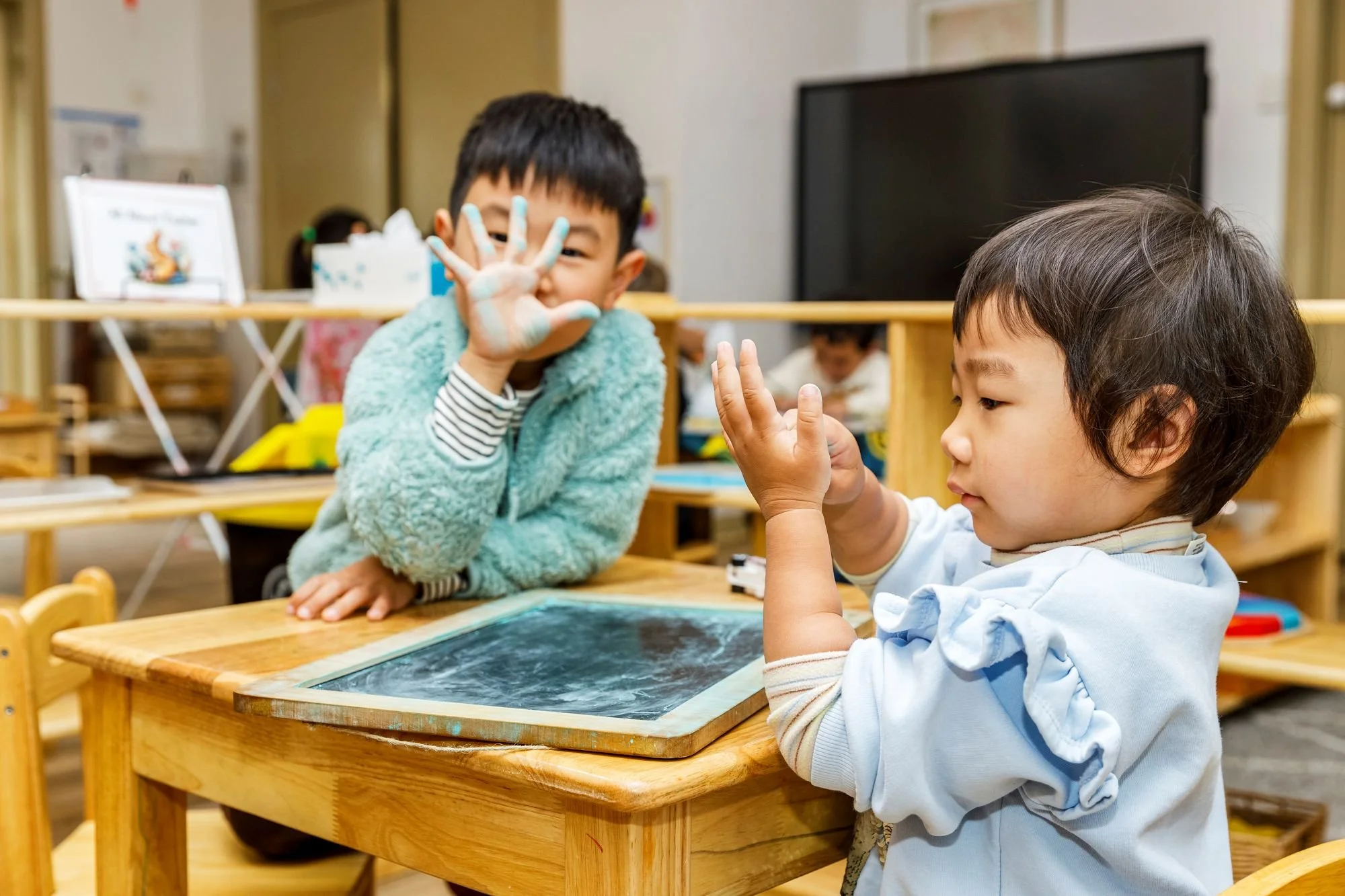 Two children, a girl and a boy, sitting at a wooden table in a classroom. The girl is holding up her hand, which is covered with writing or paint, and smiling. The boy is looking at her with interest, touching his own hand with his fingers. Other children and classroom items are visible in the background.