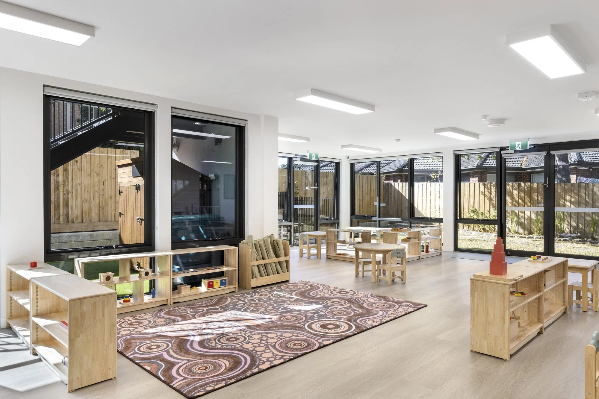 Bright, modern classroom or playroom with large windows, wooden furniture, and educational toys. The room has a patterned rug, and natural light streams in through the windows, creating a welcoming environment for children.