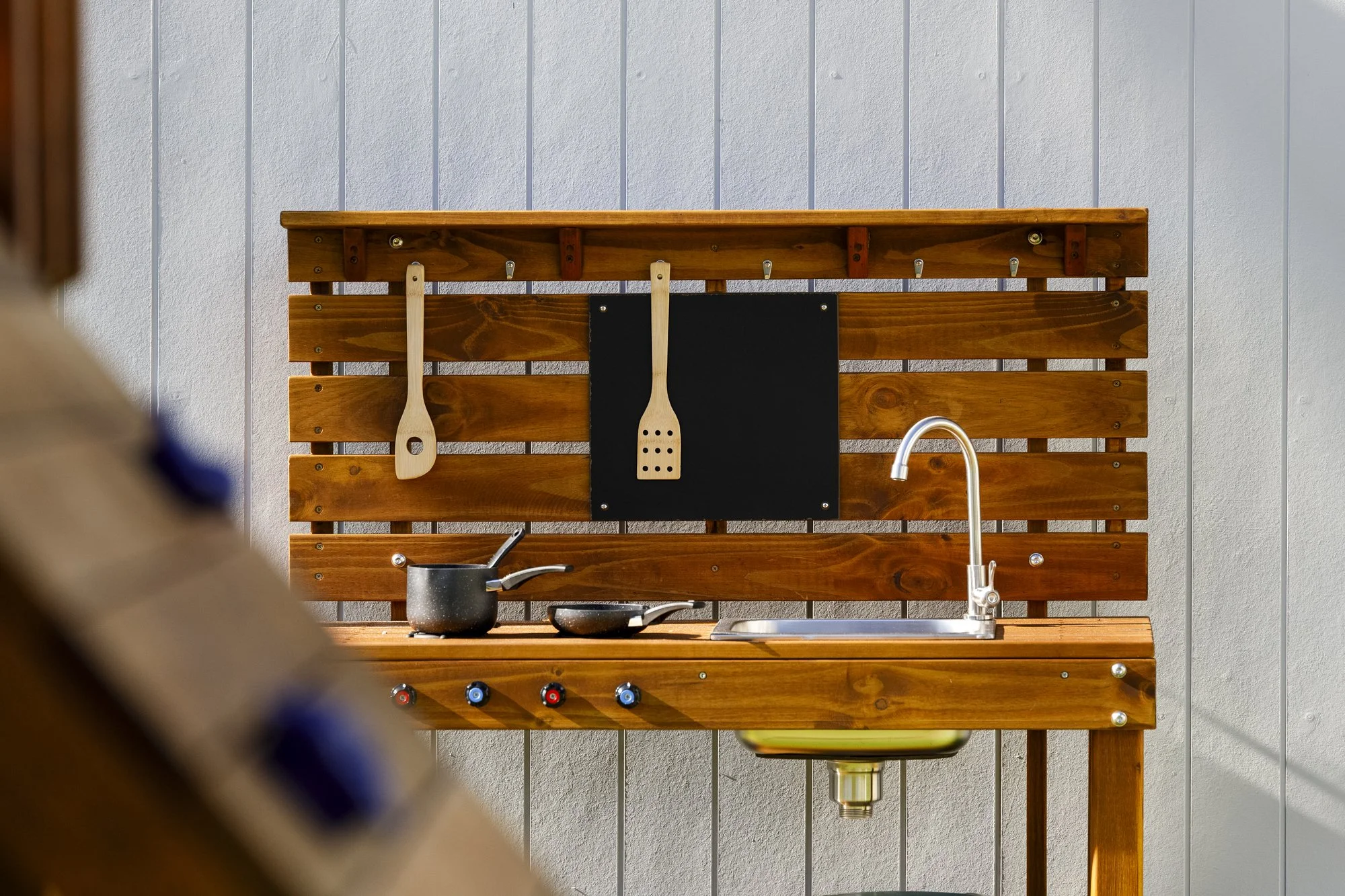 Outdoor kitchen with a wooden backsplash, pots and utensils, a black cutting board, and a sink, against a gray wall.