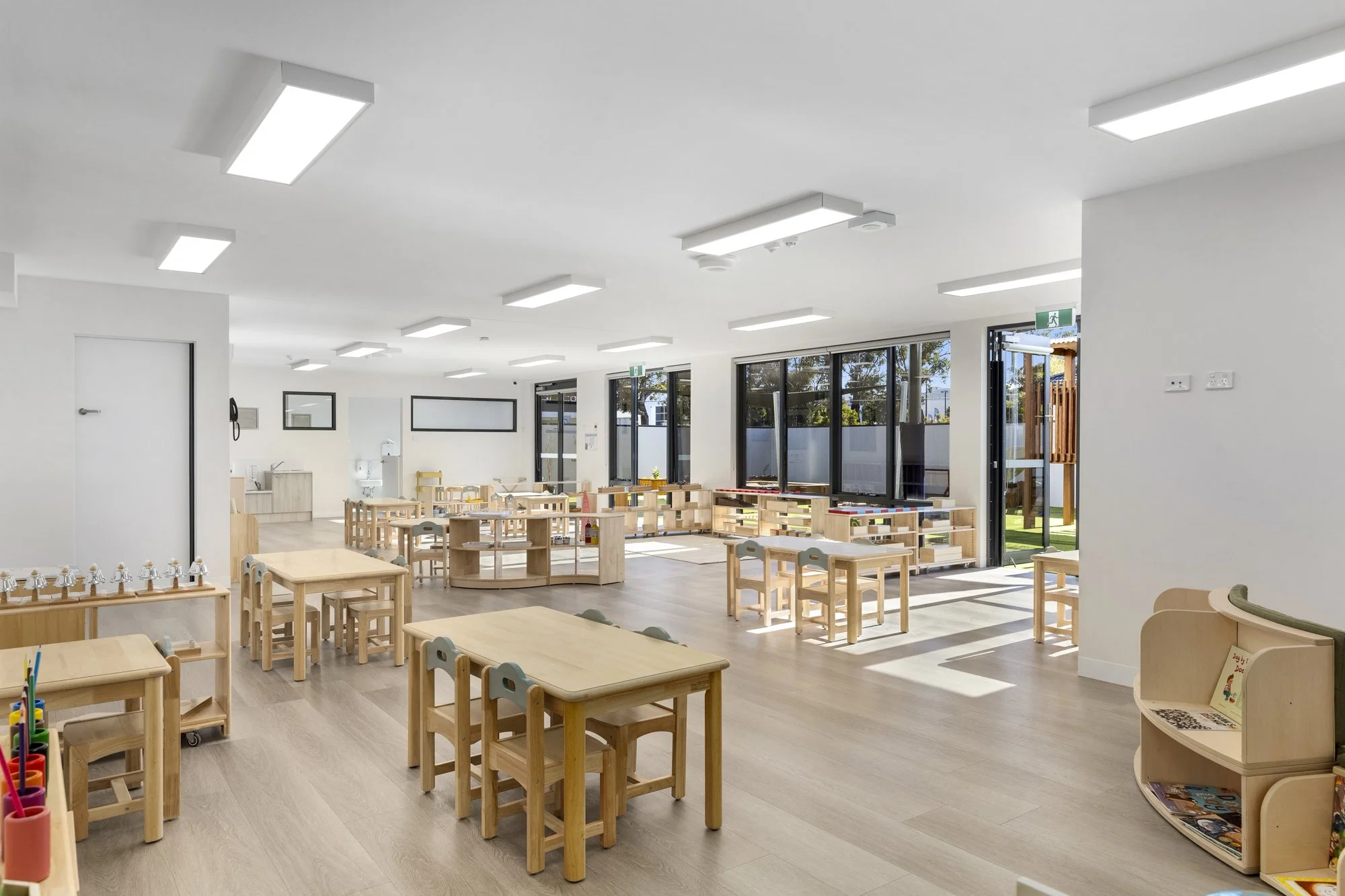 Bright, modern classroom with wooden tables and chairs, large windows letting in natural light, and educational toys and books.