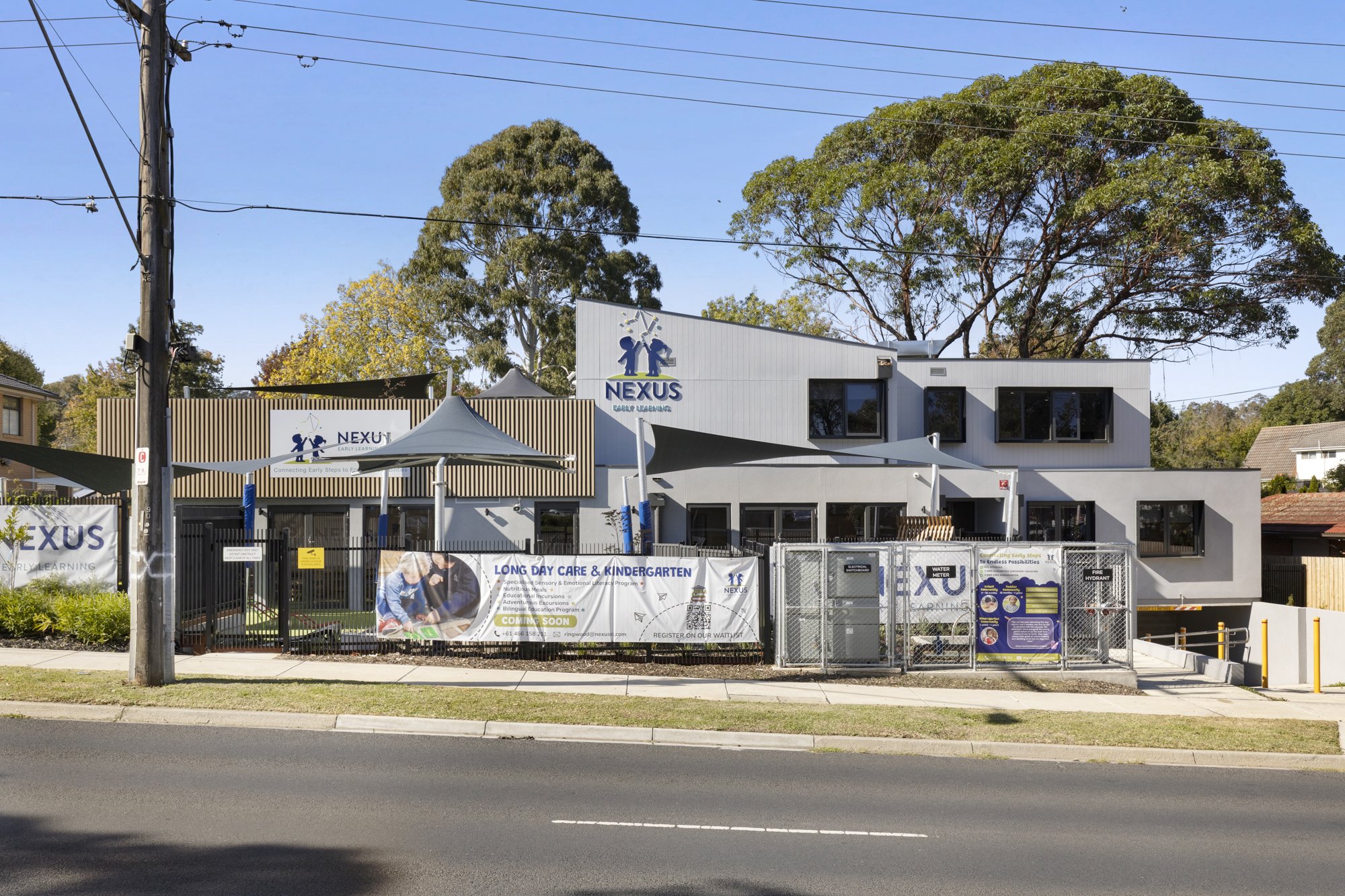 The image shows a modern building with the sign 'NEXUS Early Learning'. The building has large windows and an outdoor area with a canopy. There are trees in the background, and a utility pole with power lines is visible on the left side. A fence with