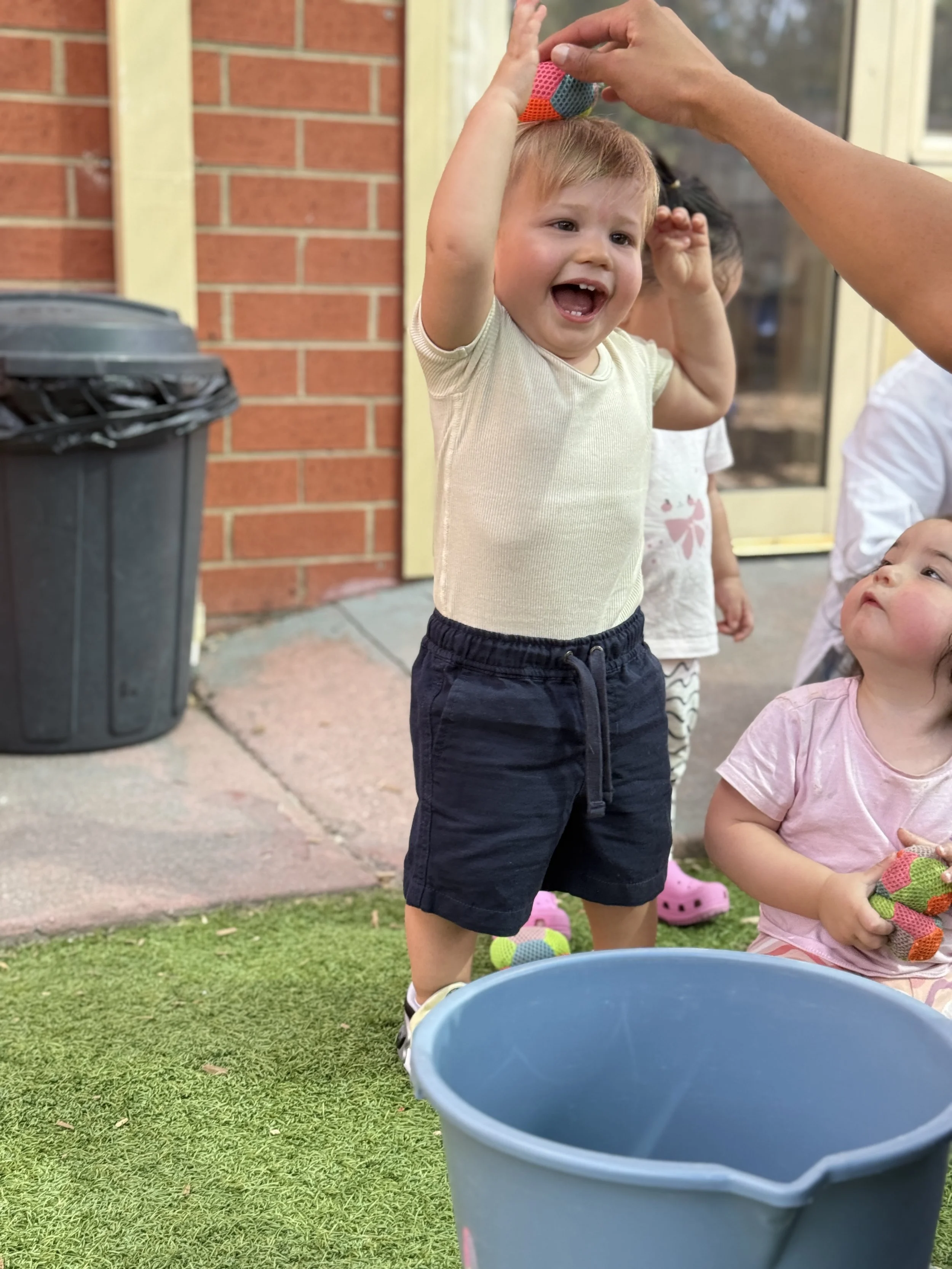 A young child with blond hair, wearing a cream-colored t-shirt and navy shorts, is reaching up towards an adult's hands holding a colorful ball. There are other children nearby, one with dark hair and a white shirt, and another with curly hair and a pink shirt, all outside near a brick house with yellow trim. A gray trash can is on the left, and a blue bucket is in the foreground.