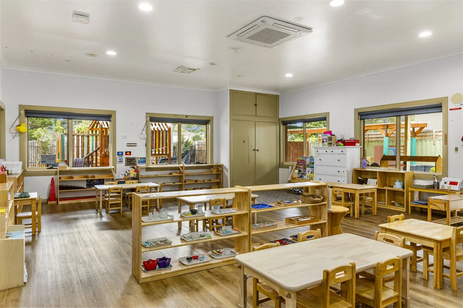 A bright classroom with wooden tables, chairs, and shelves, large windows showing outdoor play area, and educational materials and toys organized around the room.