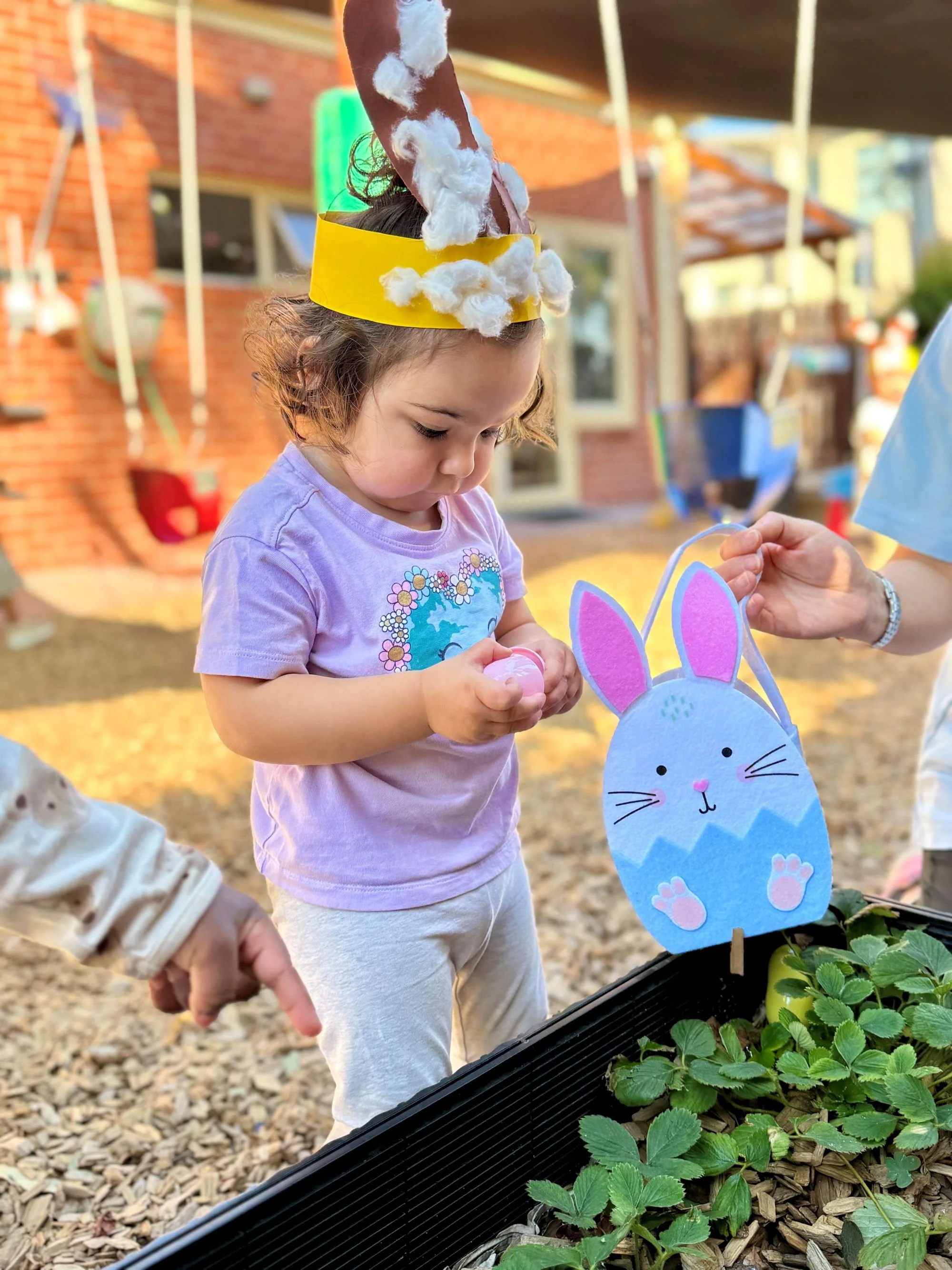 A young girl with a handmade bunny-eared hat made of paper and cotton, looking down and holding a small object, near a plant with green leaves in a garden setting.
