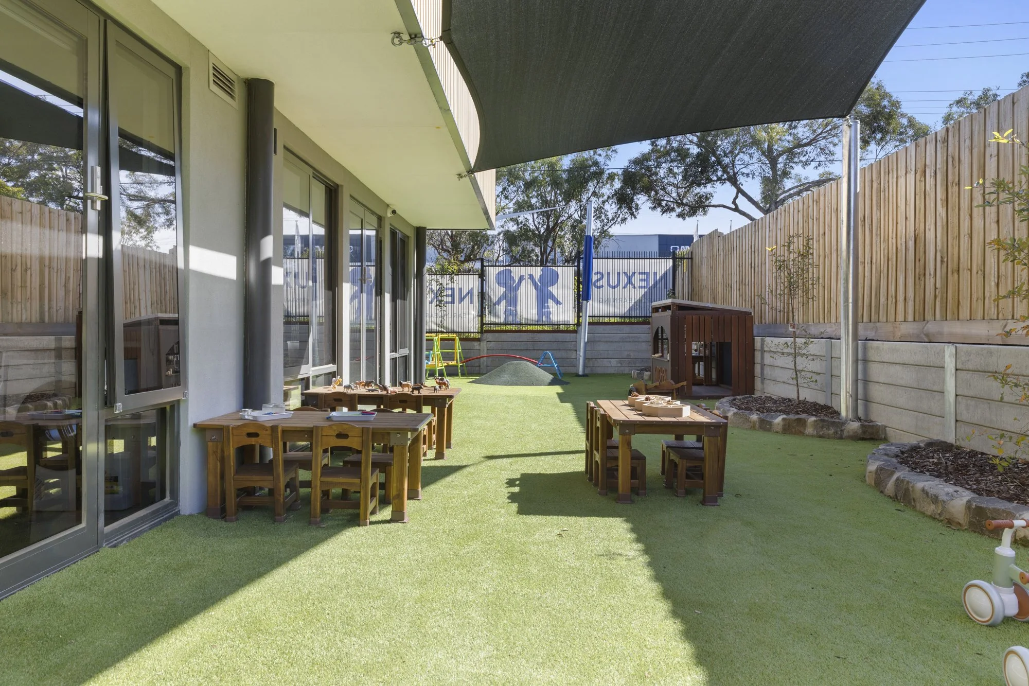Child's outdoor play area with wooden tables, toys, and playground equipment, enclosed by a wooden fence, with trees in the background.