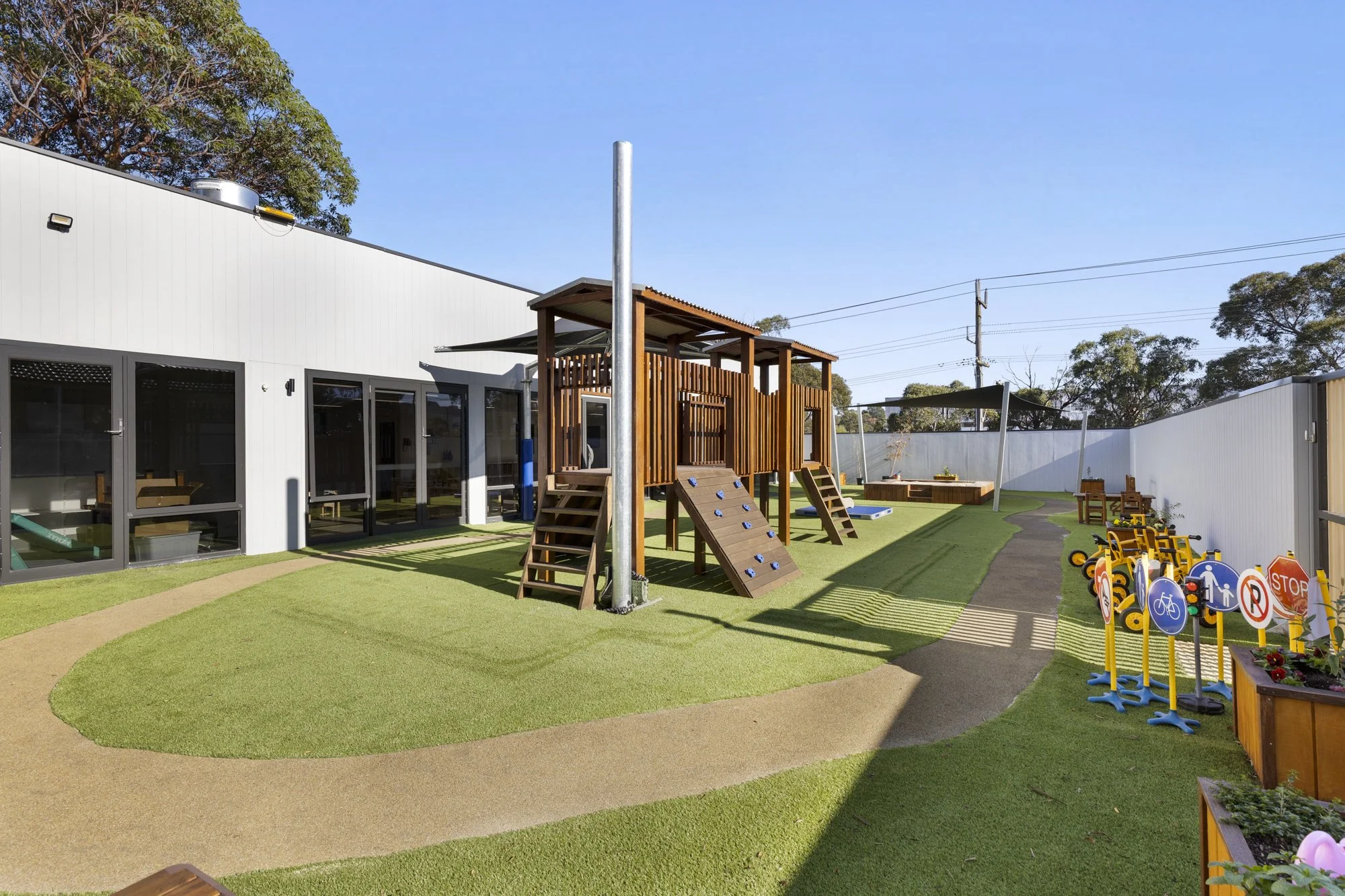 A colorful outdoor playground with a play structure, climbing wall, sandbox, and yellow ride-on toys, surrounded by a white fence.