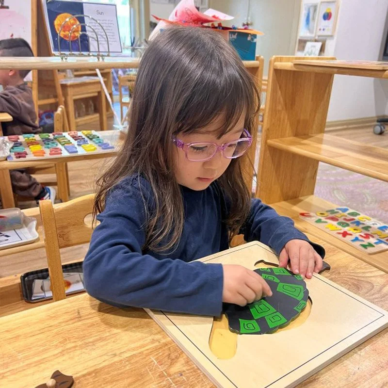 Young girl with glasses working on a shape puzzle in a classroom.