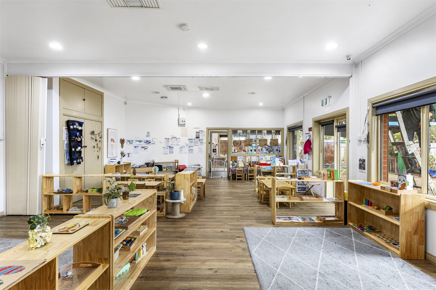 Empty classroom or playroom with wooden shelves and tables, decorated with plants and toys, large windows, and educational materials on walls.