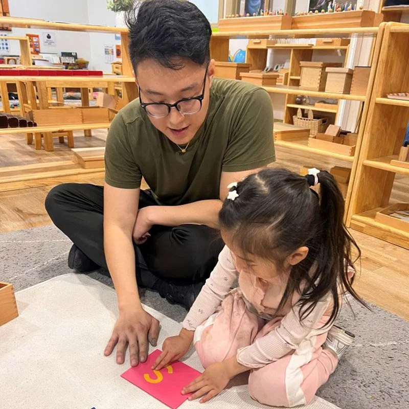 A man with glasses and a young girl are sitting on the floor in a classroom or library. They are looking at a pink card with yellow text. The girl is pointing at the card, and the man is watching her attentively.