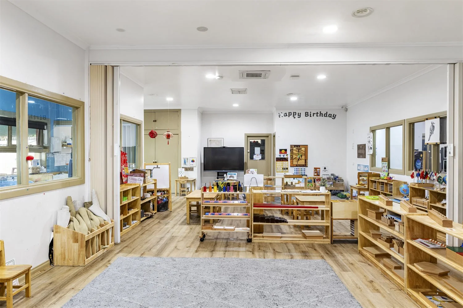 An empty classroom with wooden shelves, tables, and chairs, decorated with children's artwork and educational materials, celebrating a "Happy Birthday" with red and black decorations, bright and organized for young students.