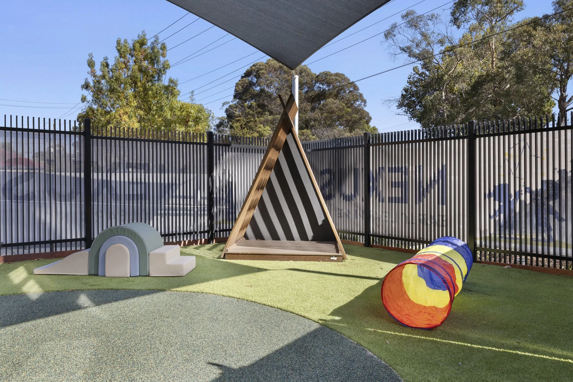 A fenced outdoor play area with a small wooden triangular structure, a rainbow tunnel, foam climbing blocks, and a slide on a synthetic grass surface, with trees and a clear blue sky in the background.