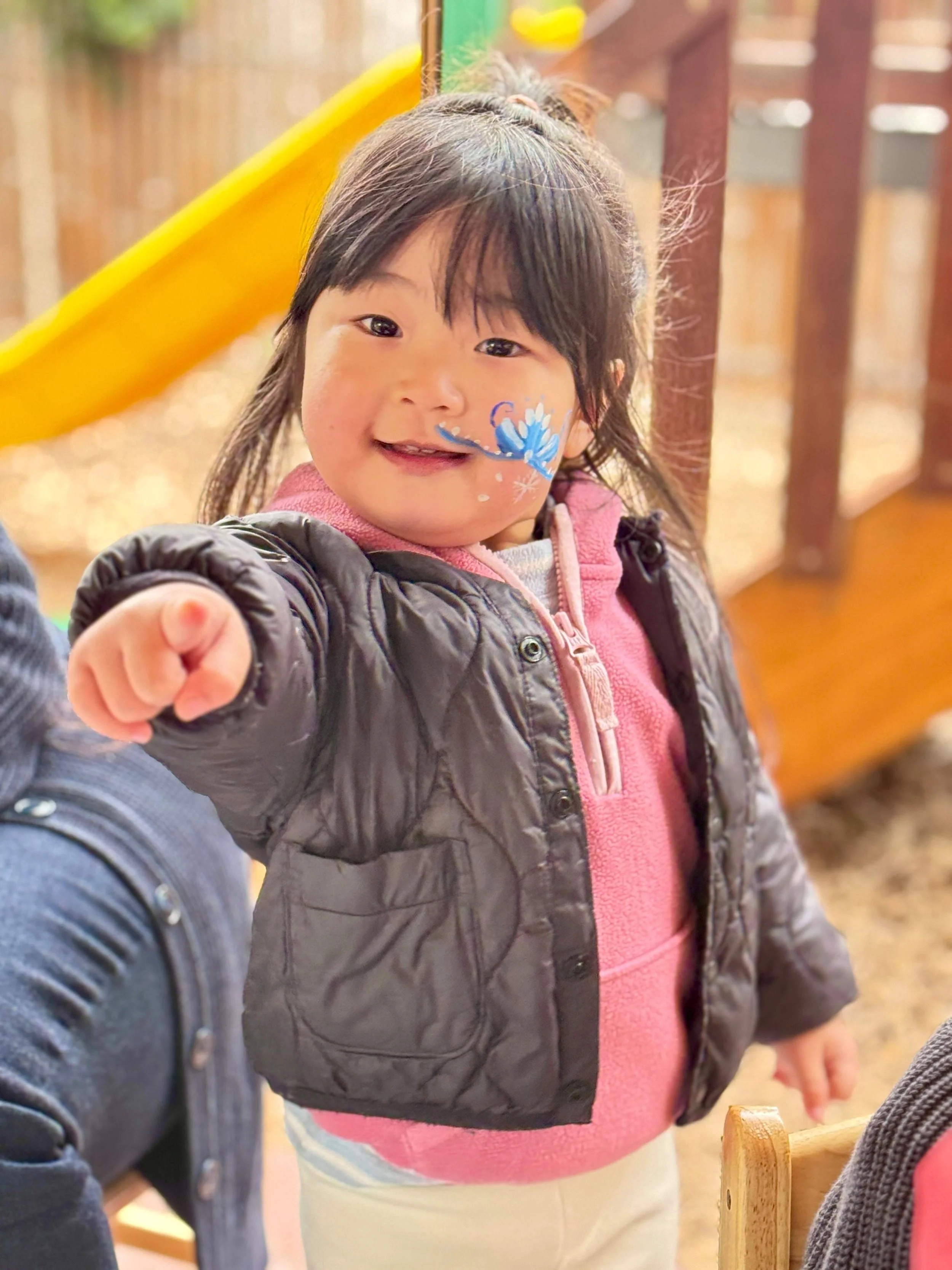 Close-up of a young girl with face paint of a blue butterfly, pointing at the camera, wearing a pink hoodie and dark jacket, with a yellow slide in the background.