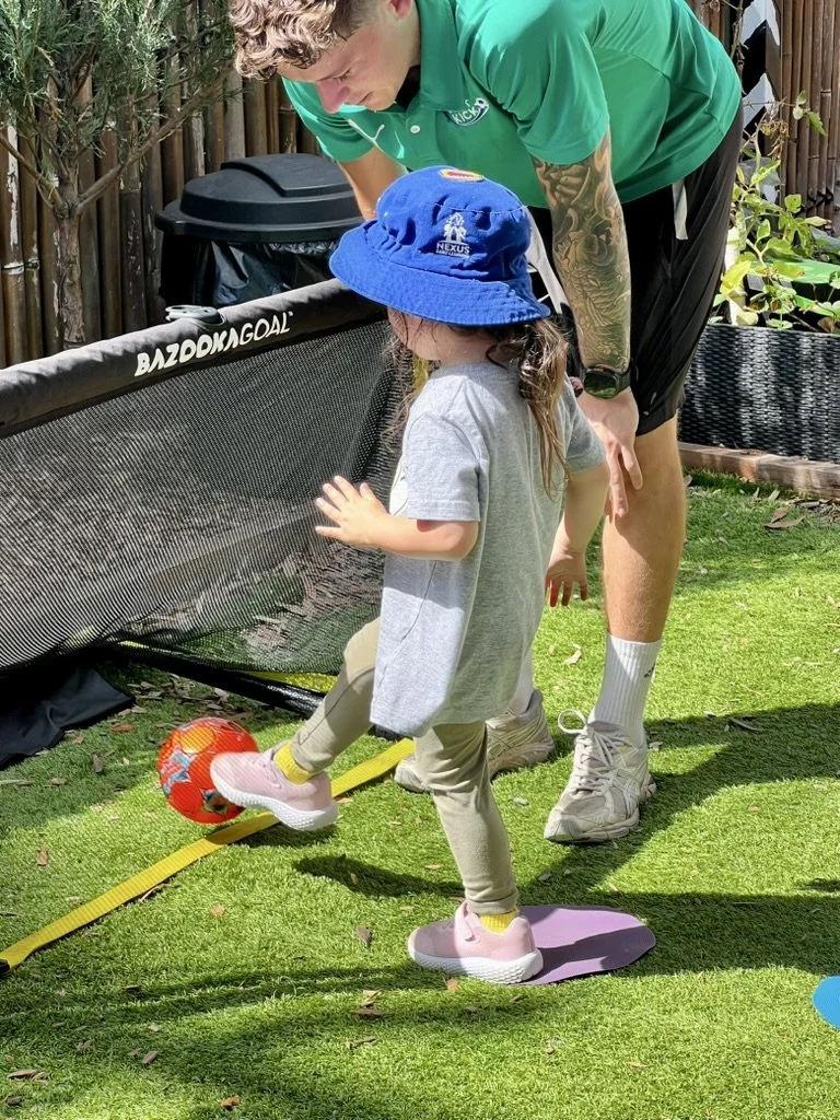 A young girl in a gray t-shirt, beige pants, pink sneakers, and a blue bucket hat plays soccer outdoors on a grassy yard. An adult male, wearing a green polo shirt, black shorts, and white sneakers, guides her as she kicks an orange soccer ball with a colorful design. There is a black mesh barrier, garden plants, and a wooden fence in the background.