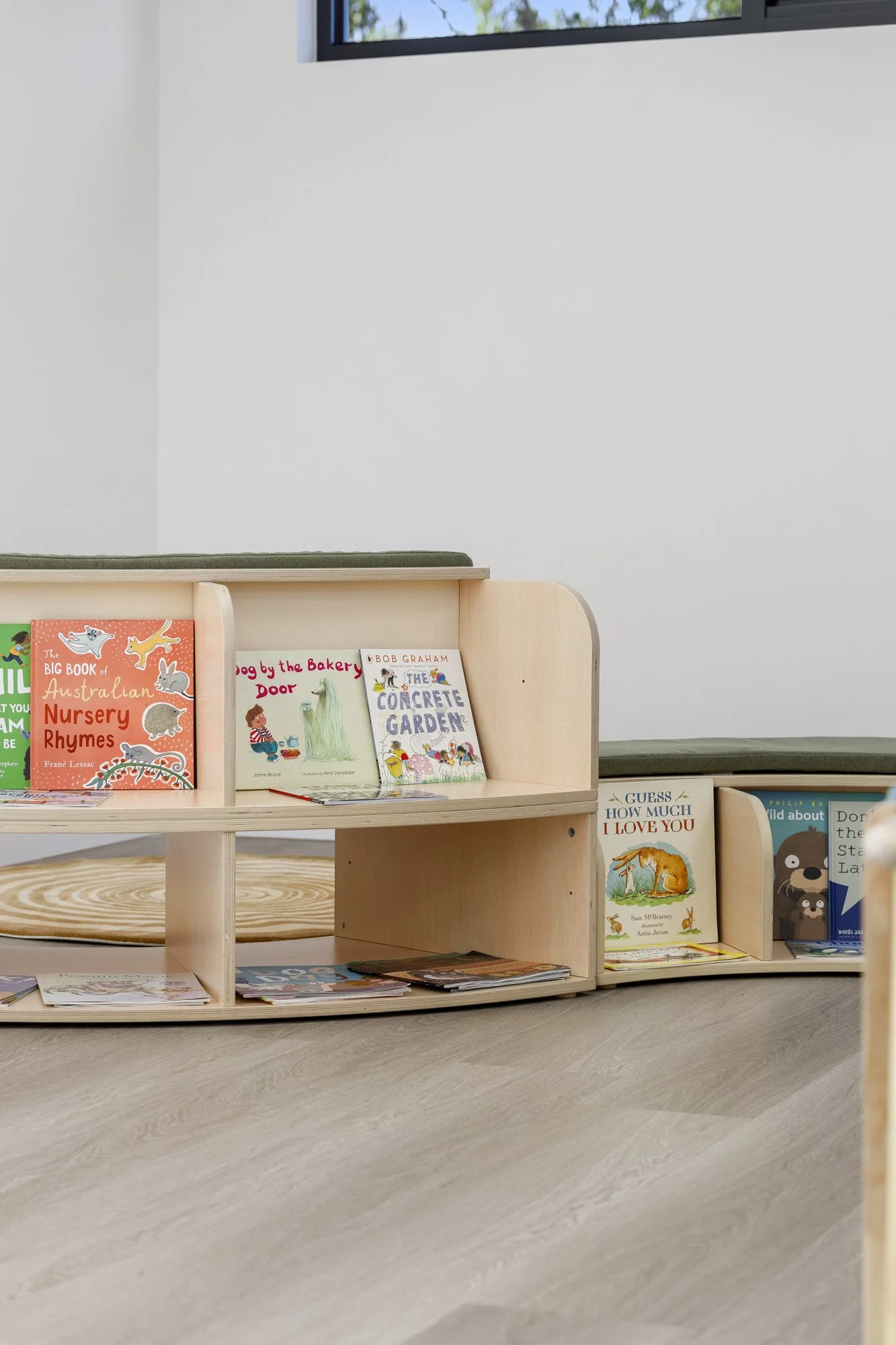 Books displayed on a wooden shelf, including titles about Australian nursery rhymes, dog stories, and animals, in a room with a window and light-colored flooring.