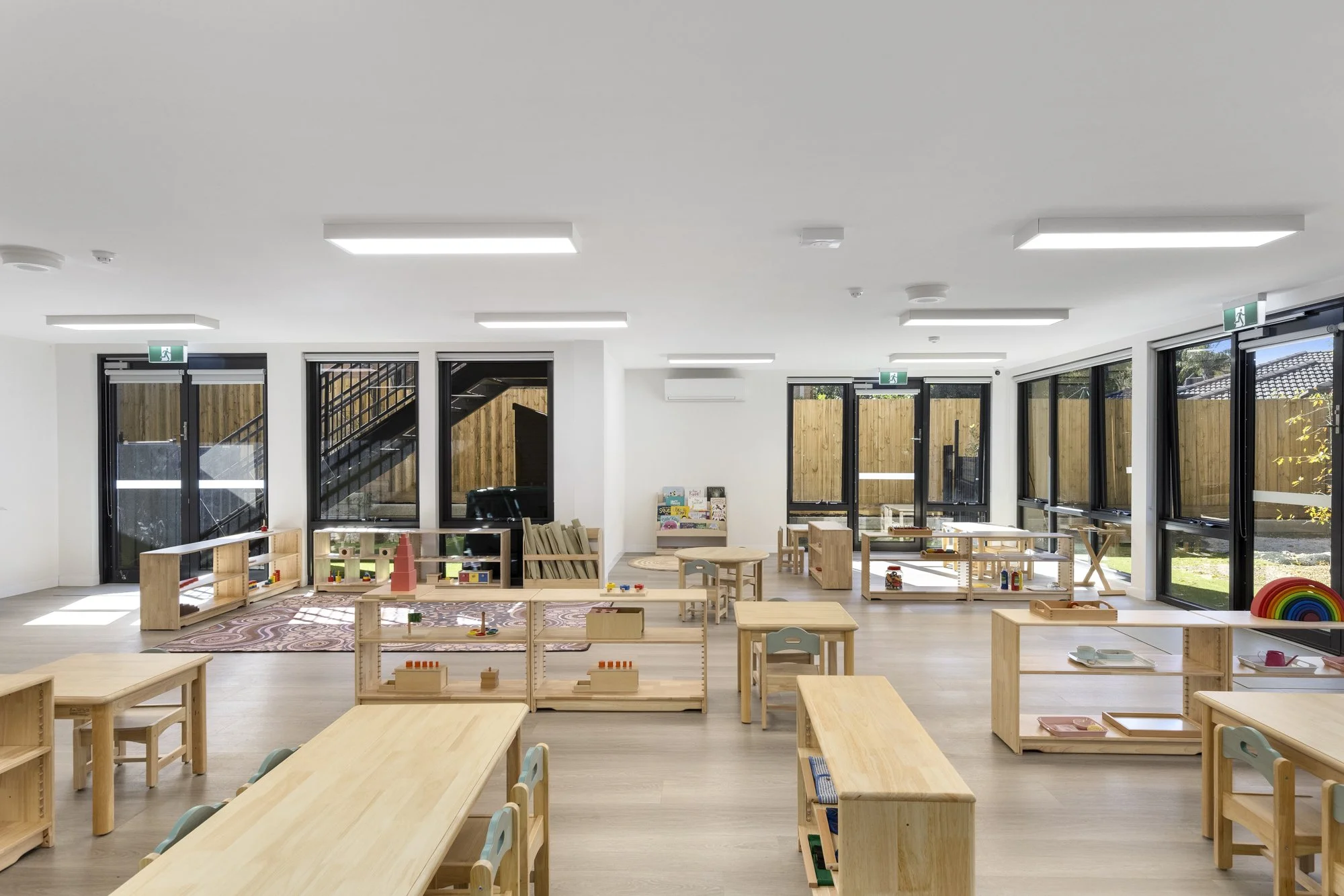 Bright, modern indoor children's play area with wooden furniture, toys, and large windows allowing sunlight, featuring exit signs and outdoor view.