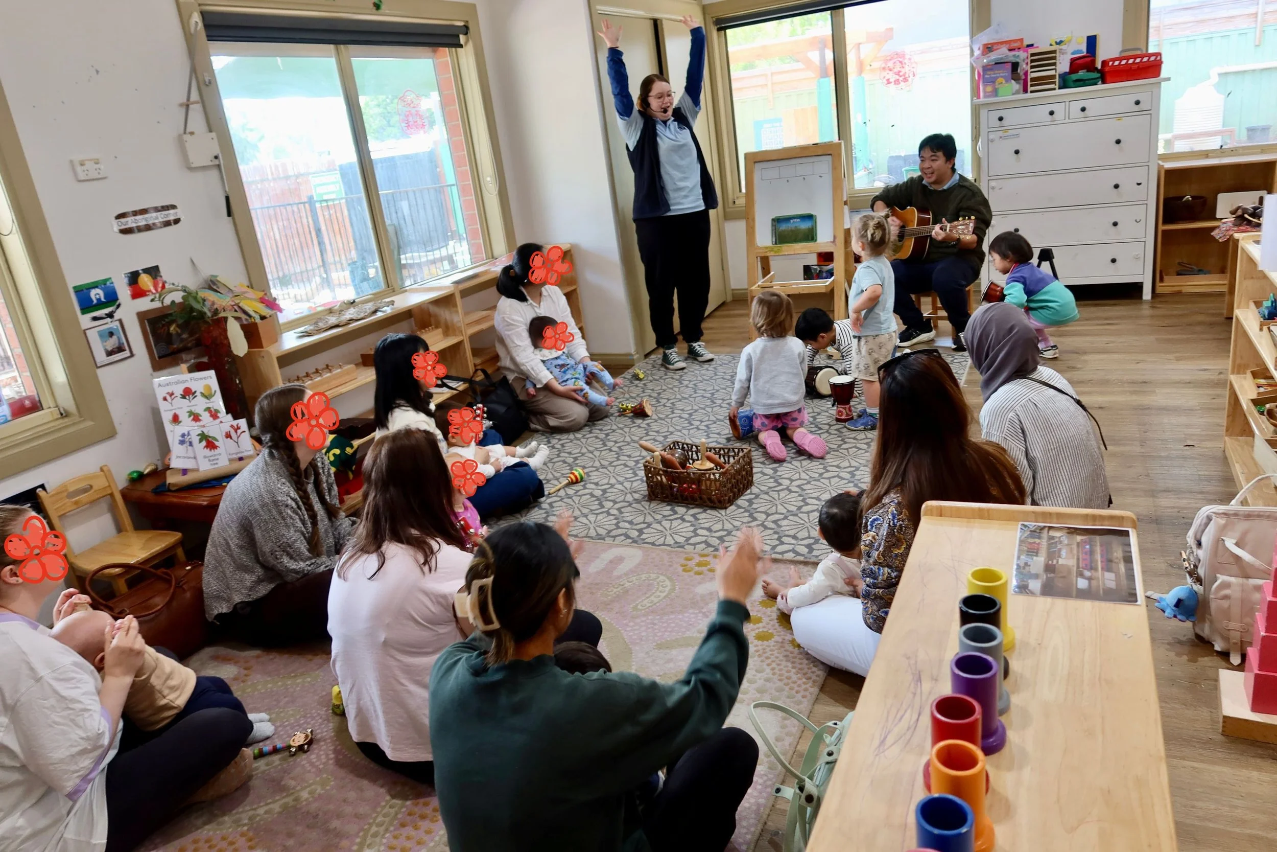 Children and adults participating in a classroom activity with two performers, one playing guitar and the other leading song or dance, while children sit on the floor and watch.