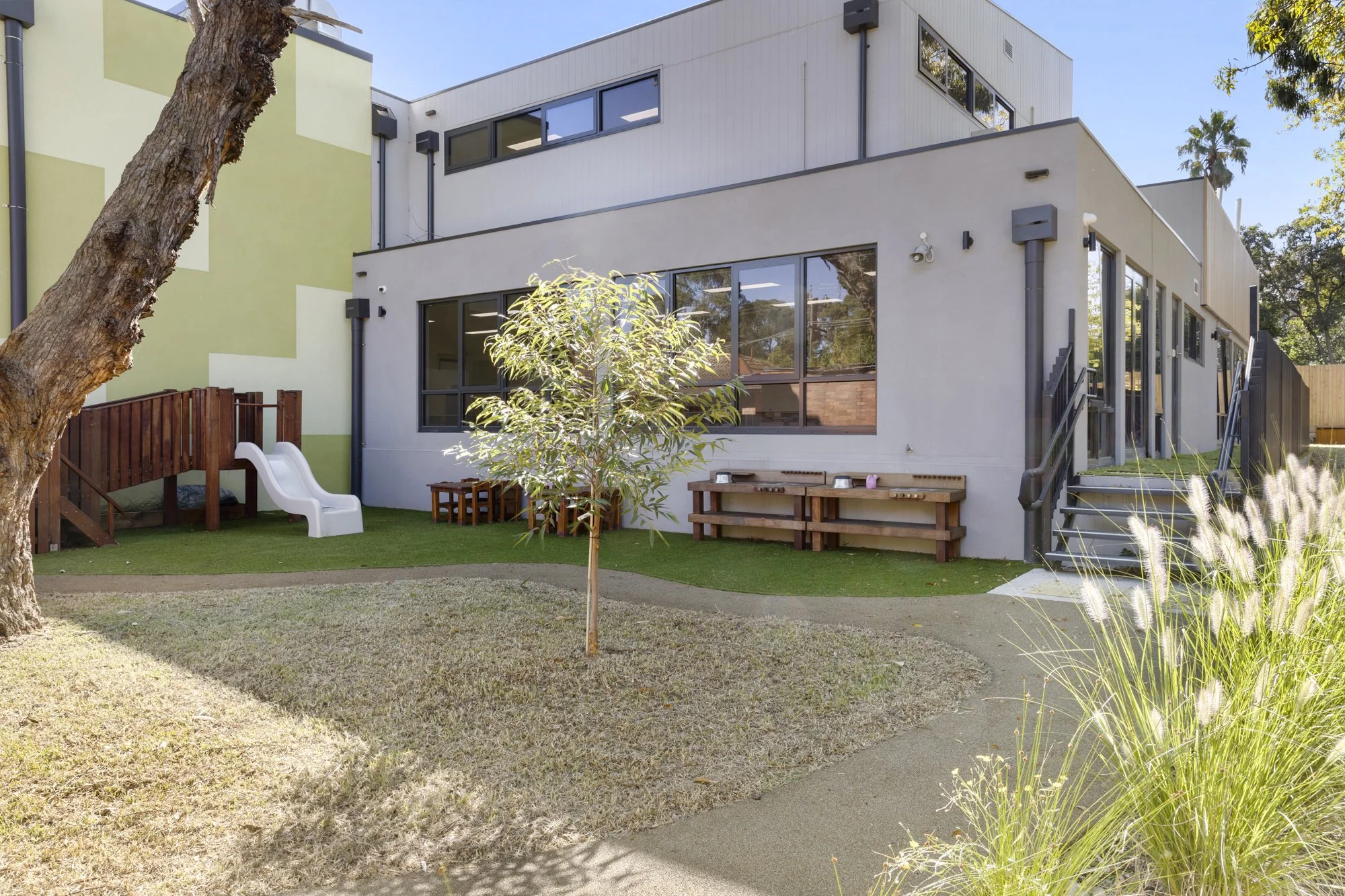 A modern multi-story building with large windows, a small outdoor yard with grass, young trees, and wooden benches. There's also a small playground slide and stairs leading to an entrance.