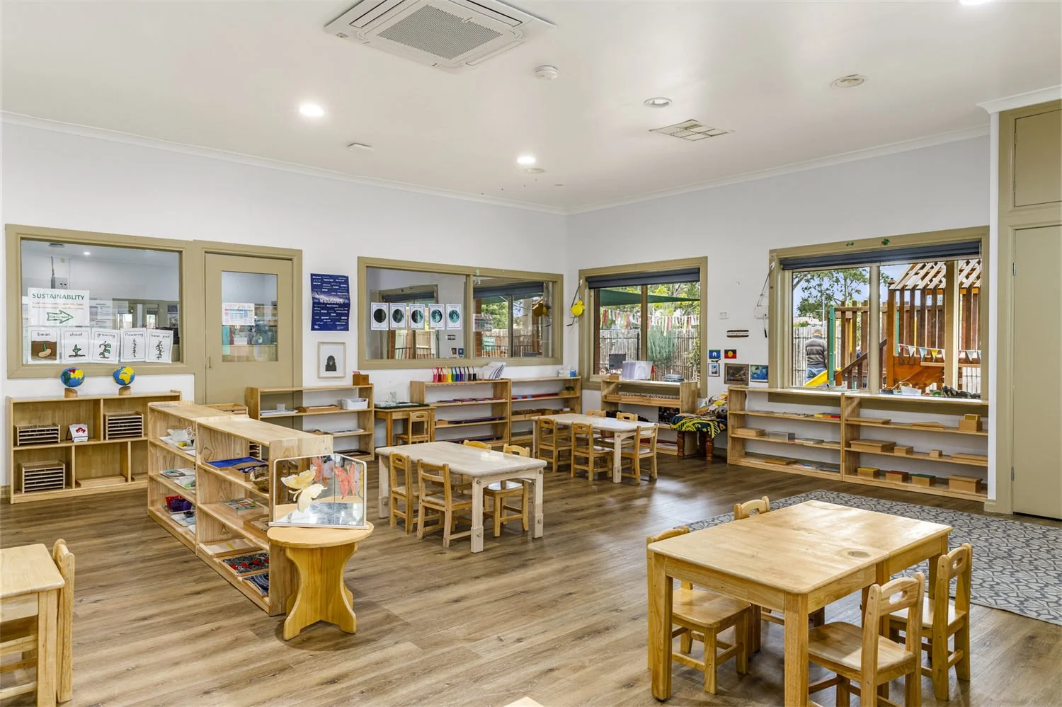 Bright and spacious preschool classroom with wooden tables and chairs, educational materials on shelves, and large windows revealing a playground outside.