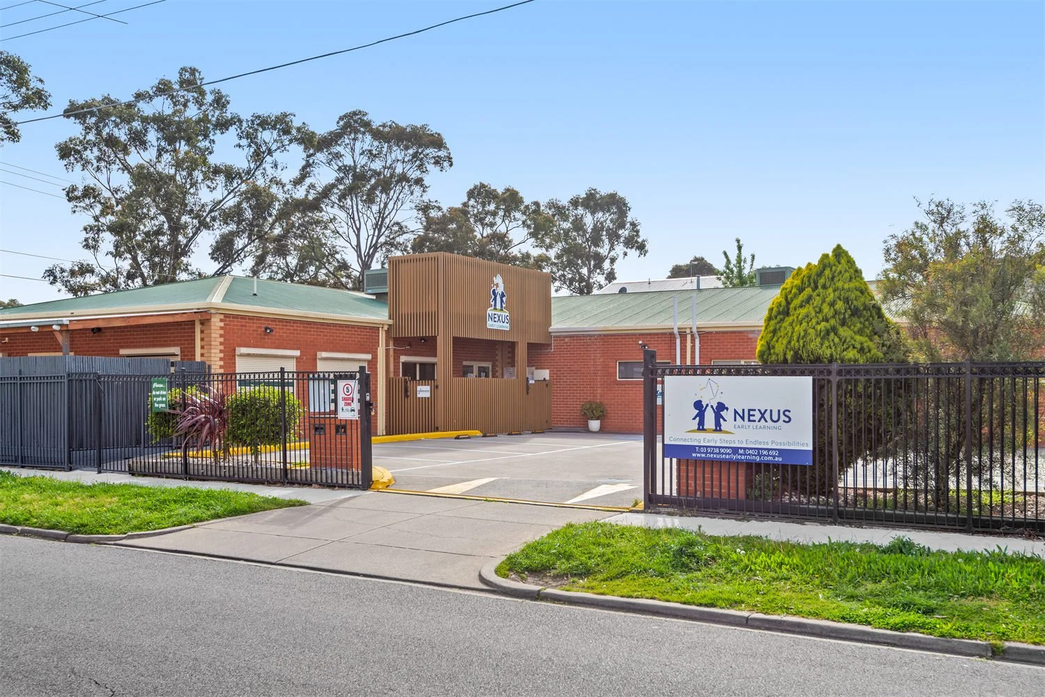 Exterior view of Nexus Early Learning preschool with a brick building, wooden entrance, and a black metal fence. Signage displays the Nexus Early Learning logo and contact details. The area has a sidewalk, some greenery, and tall trees in the backgro